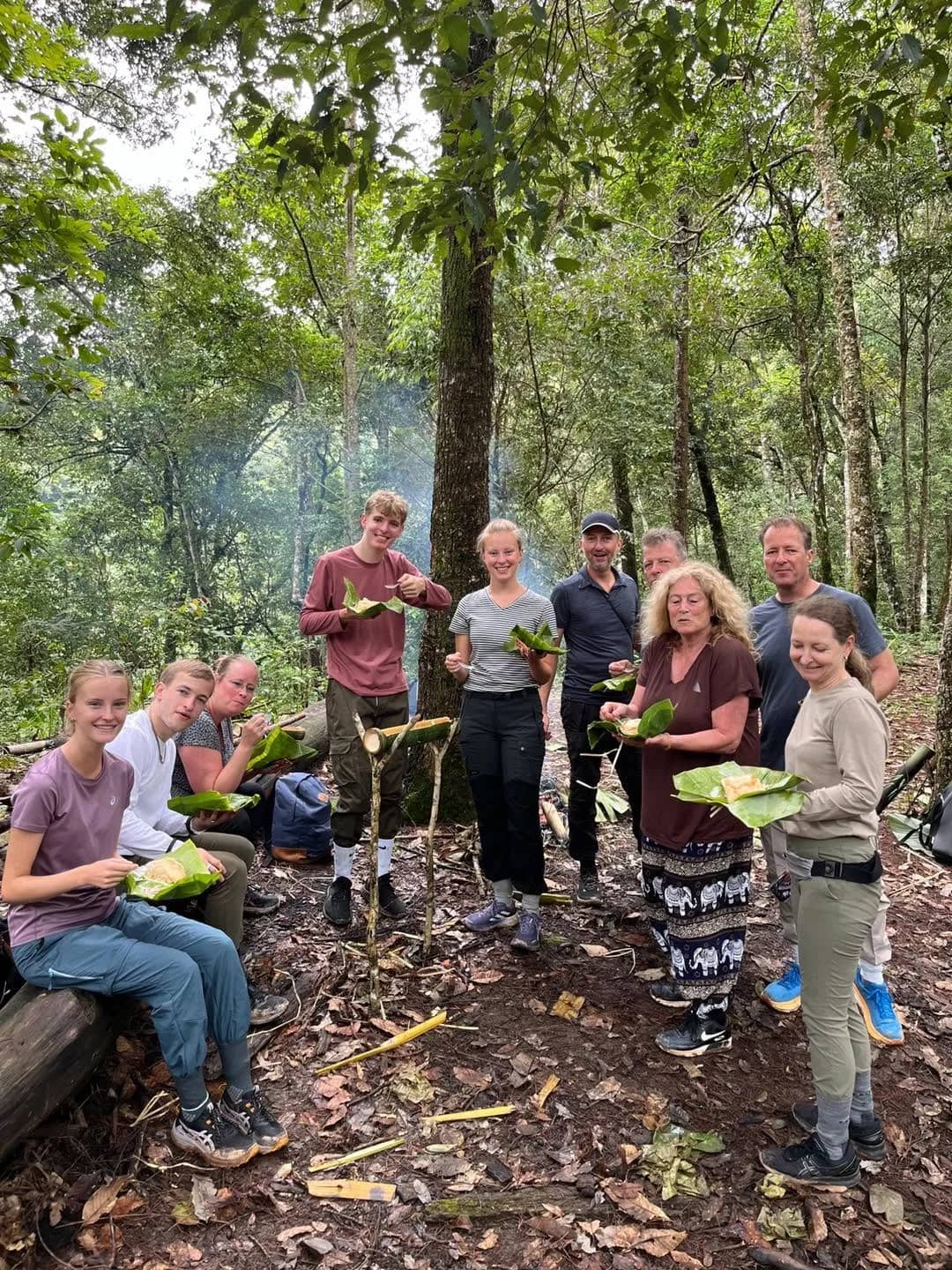 A group of trekkers holding banana leaves gathers in a forest clearing beside a smoky campfire.