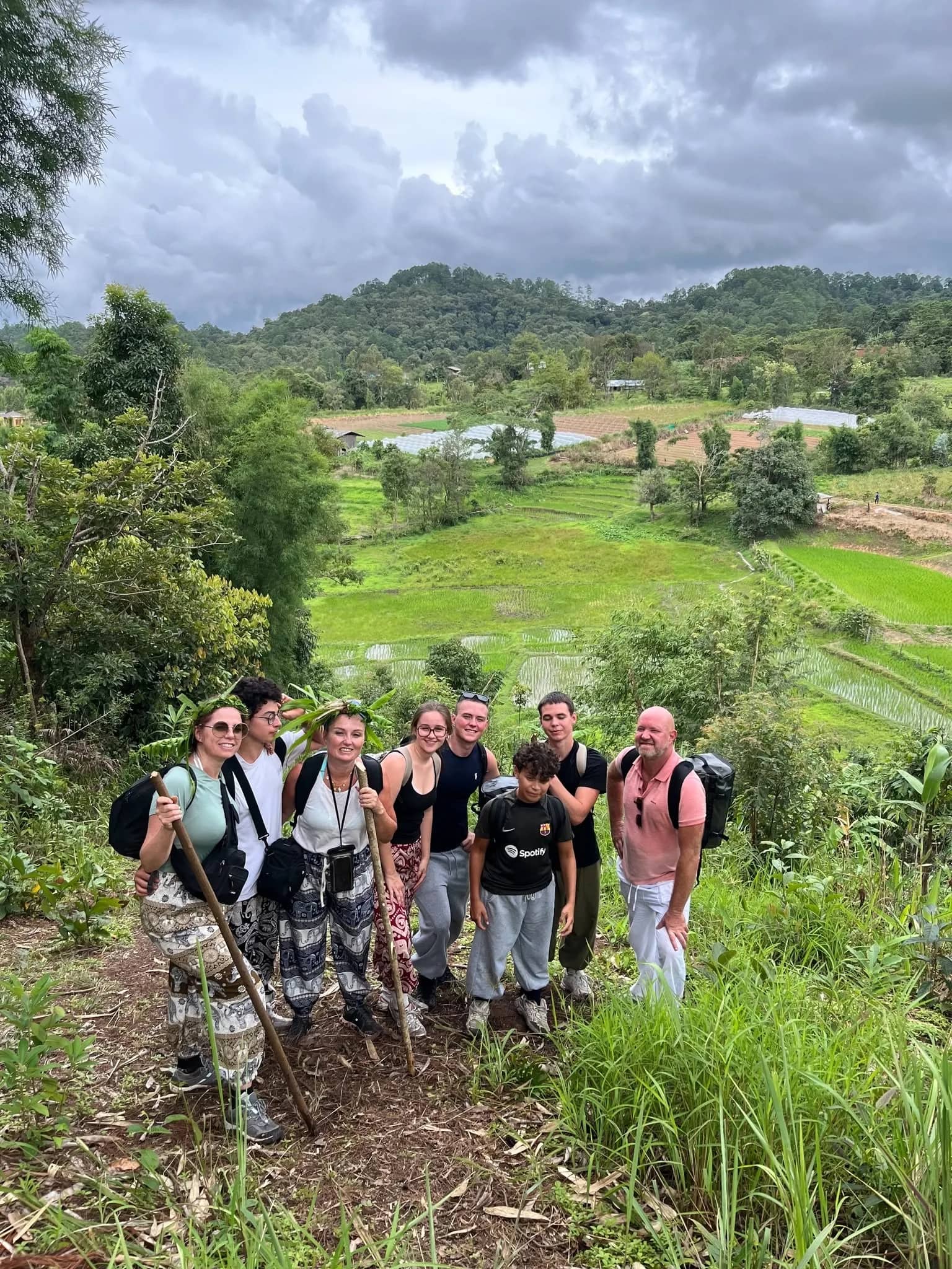 A group of trekkers with walking sticks poses on a hillside above green rice fields.