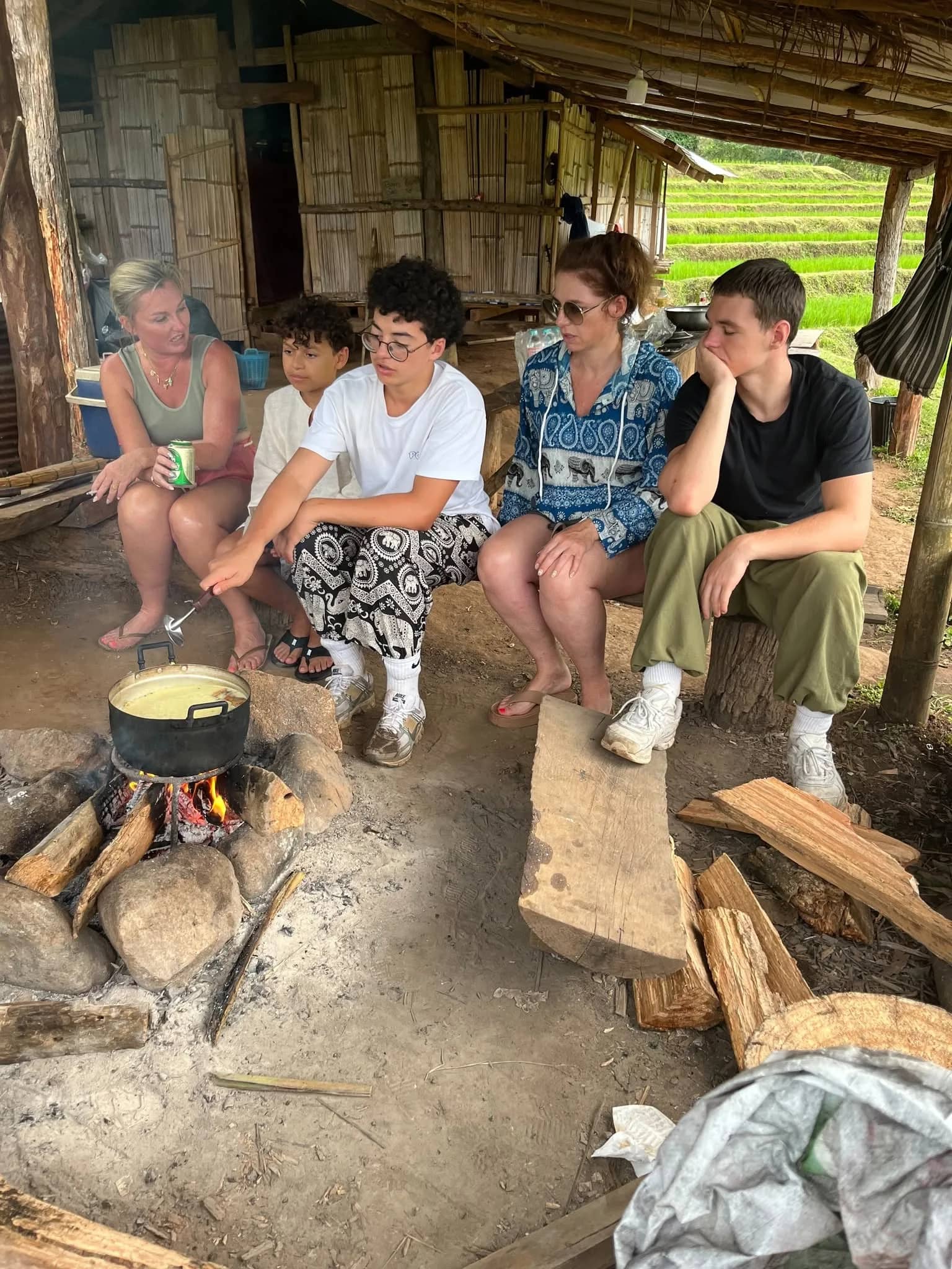 Trekkers sit around a pot cooking over an open wood fire under a bamboo hut roof.