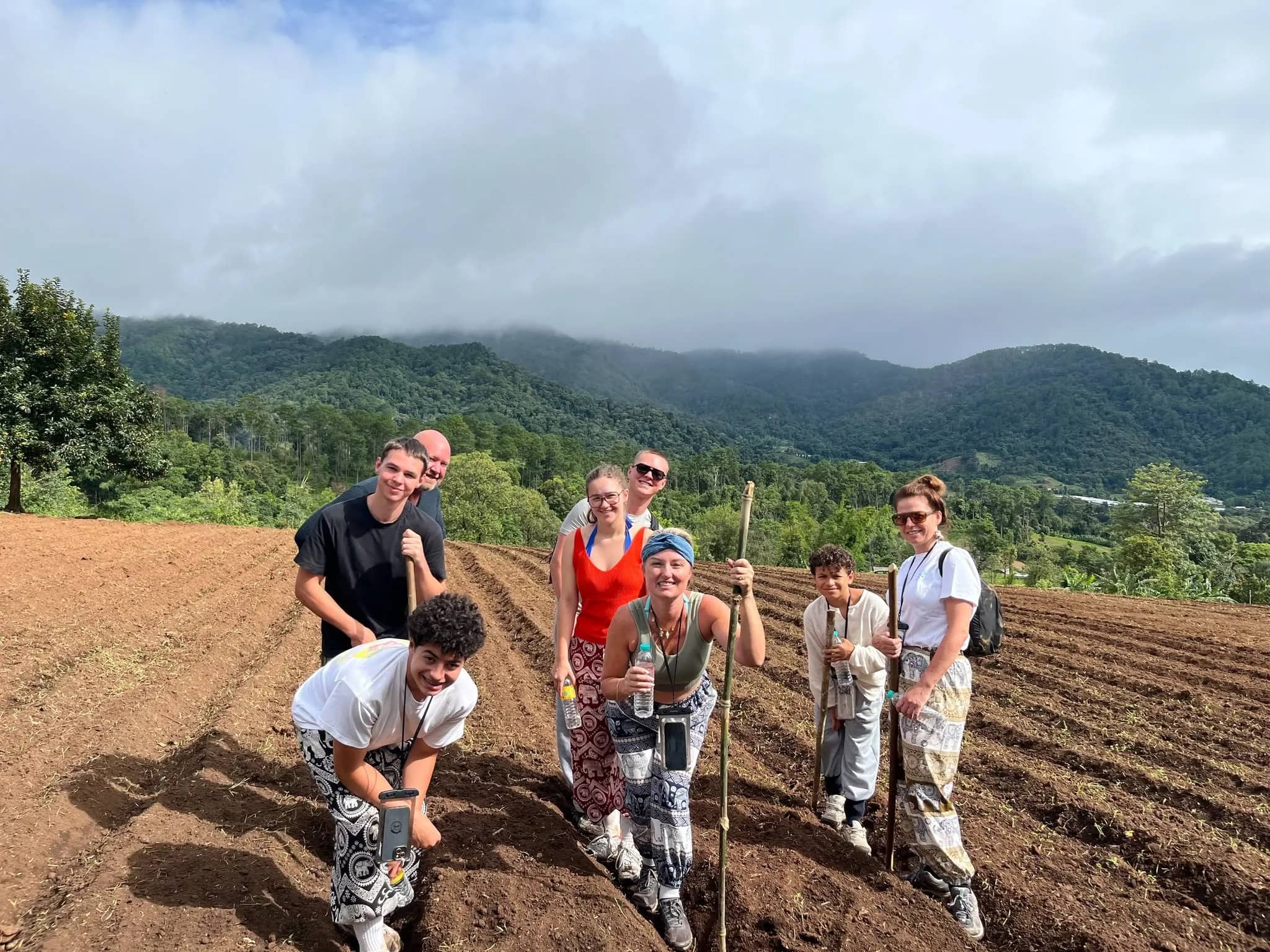 Smiling trekkers with bamboo sticks pose in a freshly plowed field with misty mountains behind.