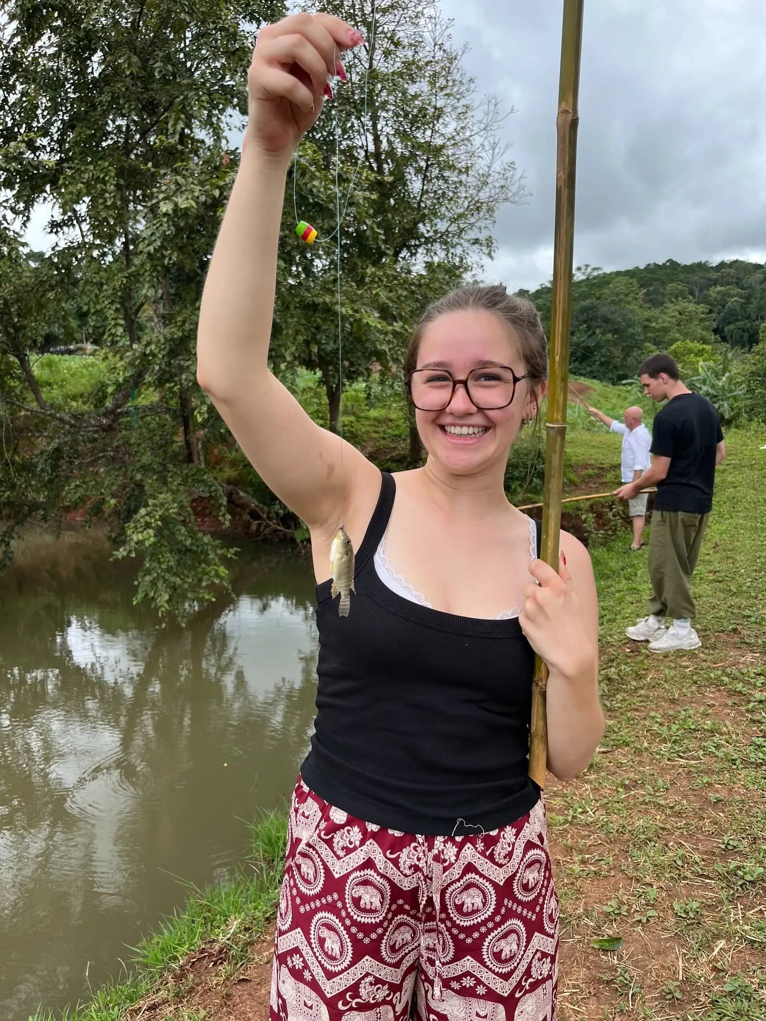 A smiling trekker holds up a small catch on a bamboo fishing pole beside a pond.
