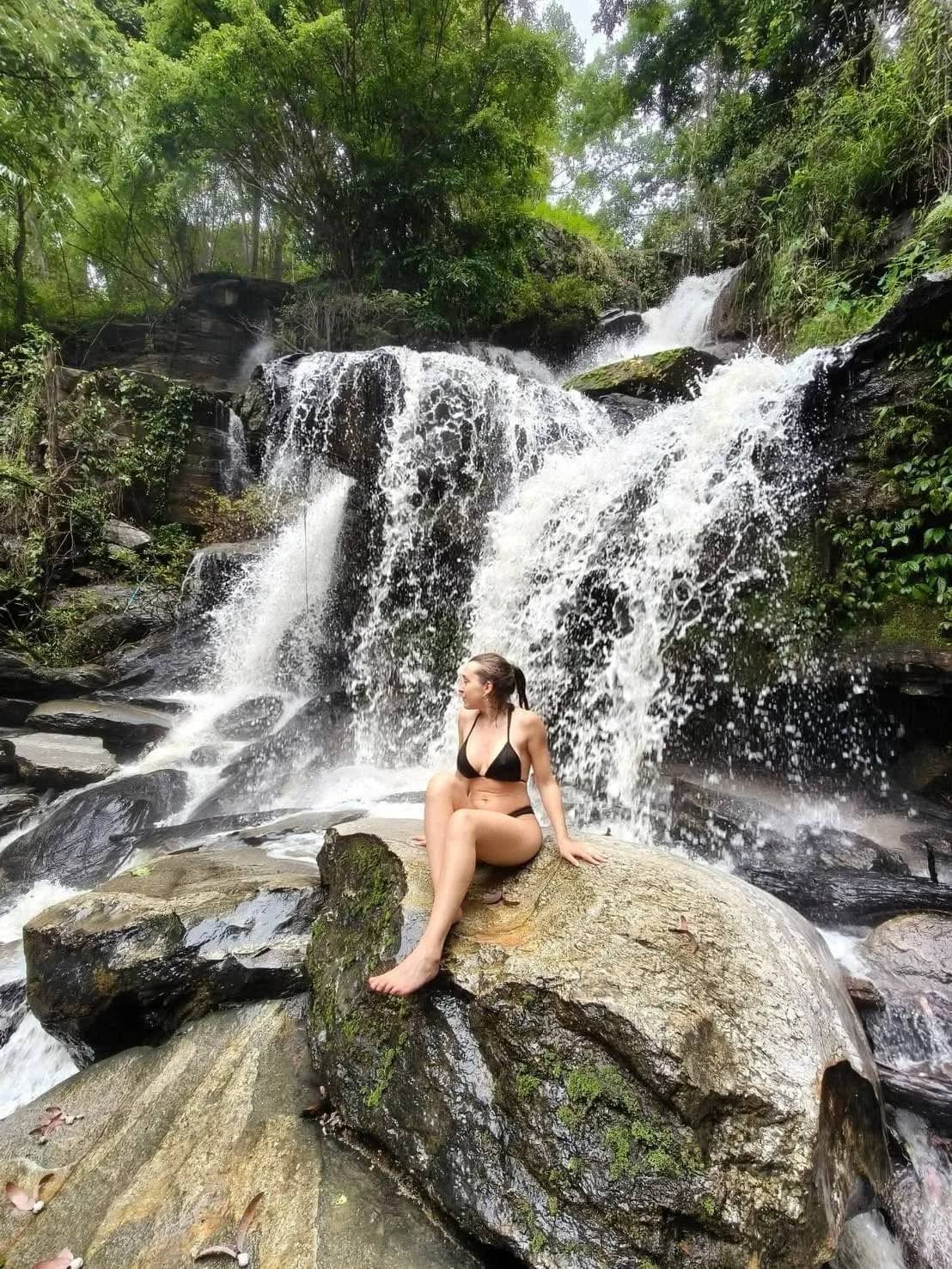 A hiker in swimwear sits on a boulder in front of a tiered jungle waterfall on the eco trek