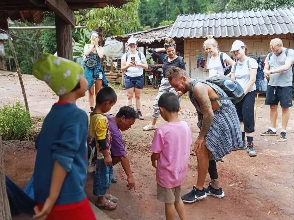Trekkers with backpacks greet local children on a dirt path in a hill village