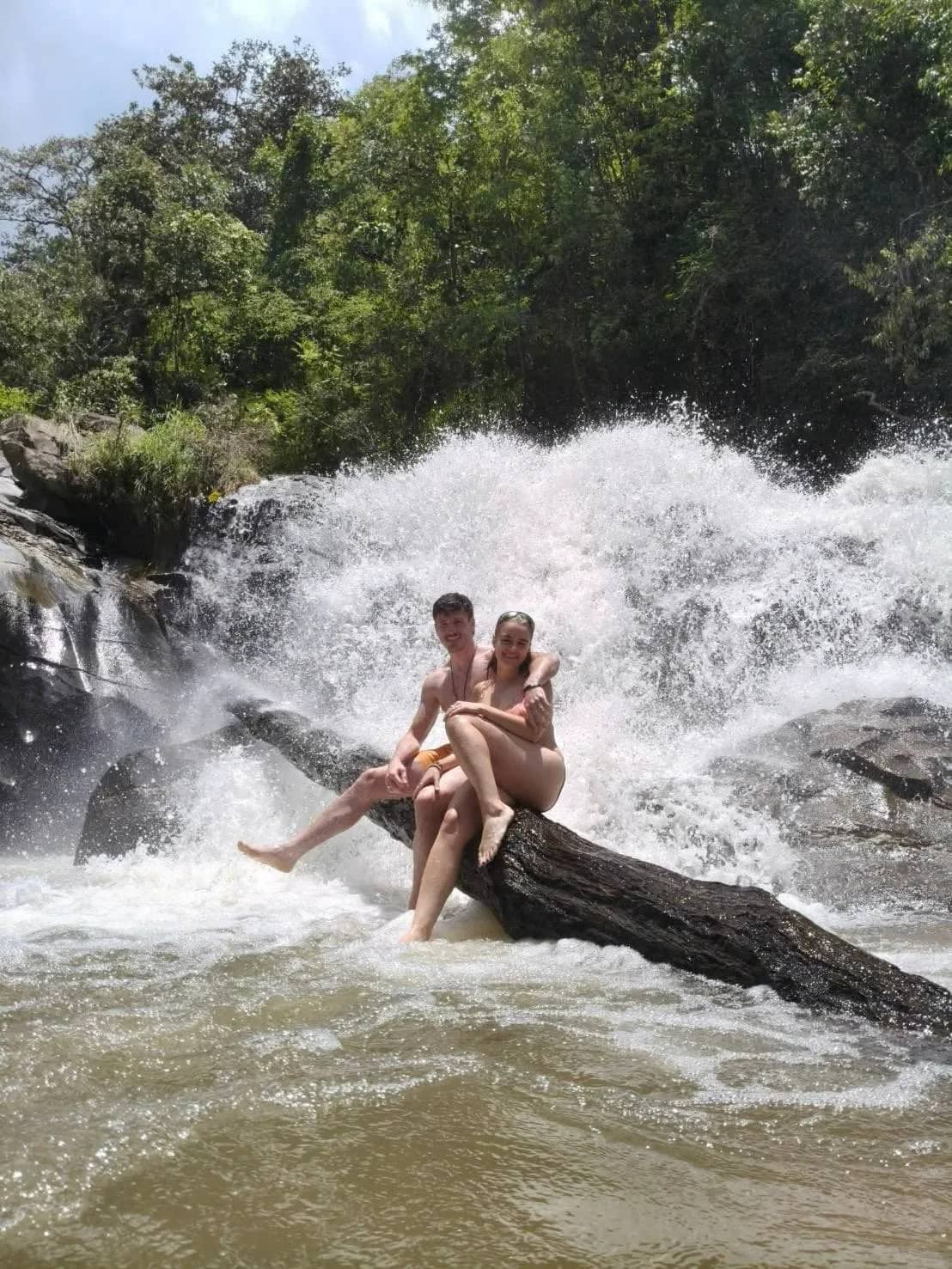 Two trekkers sit on a fallen log in the white spray of a powerful waterfall