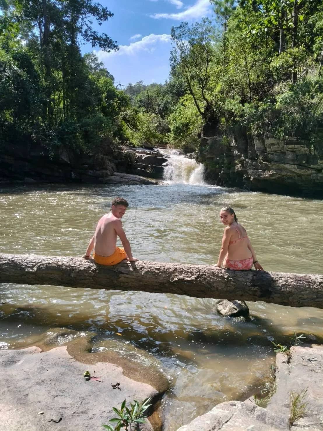Two trekkers sit on a log across a clear stream with a small waterfall behind them