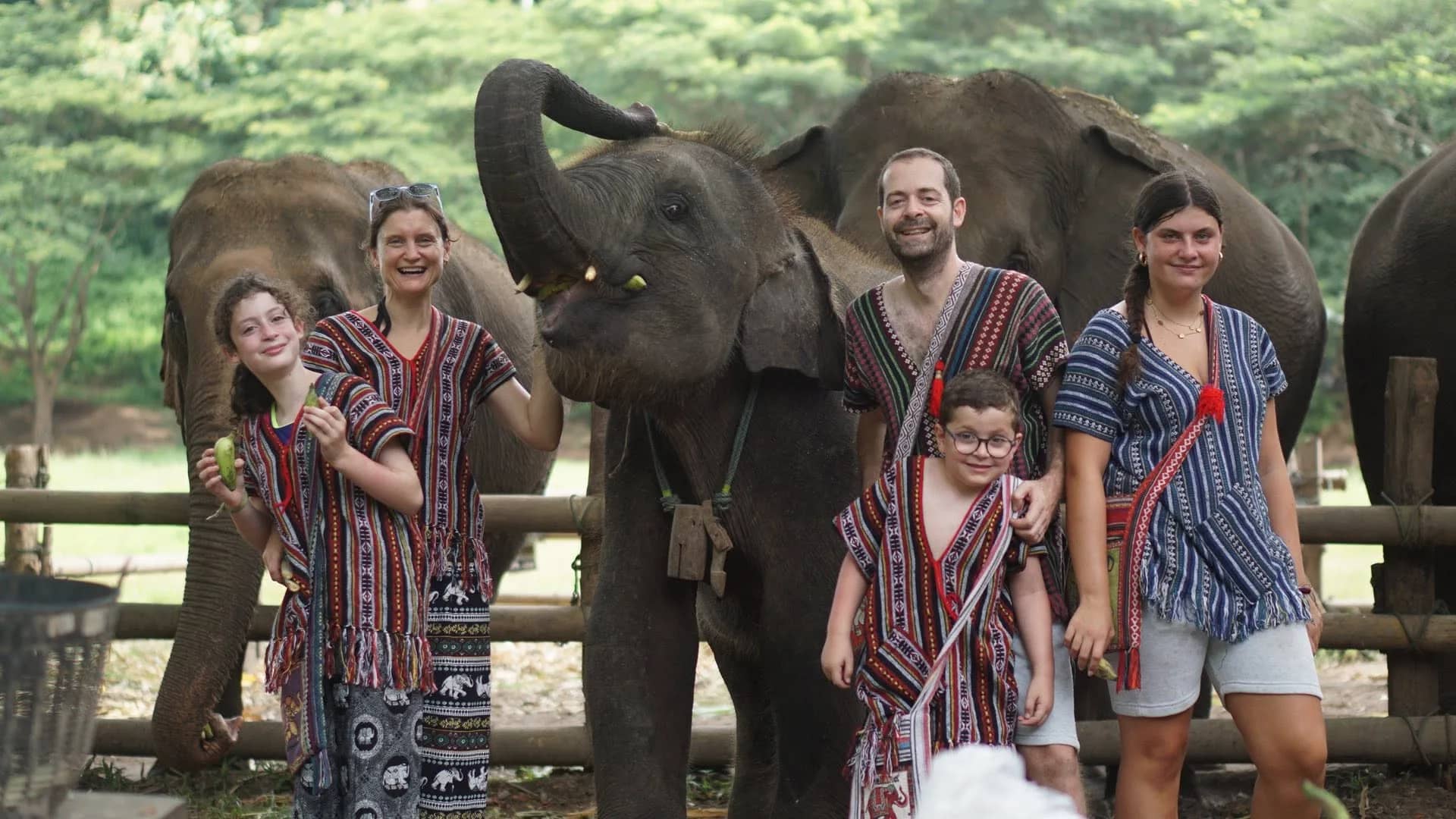 A family in traditional woven tops smiles beside elephants in front of a wooden fence.