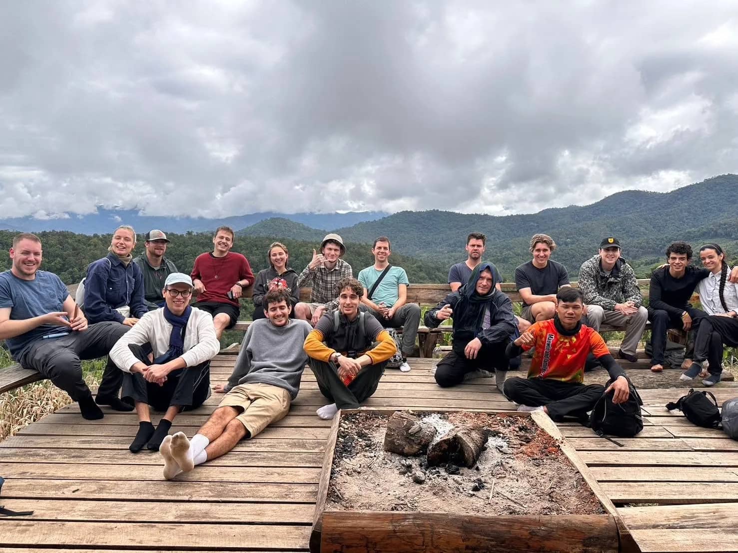 Trekkers sit around a fire pit on a wooden deck with cloudy mountain views at Doi Inthanon