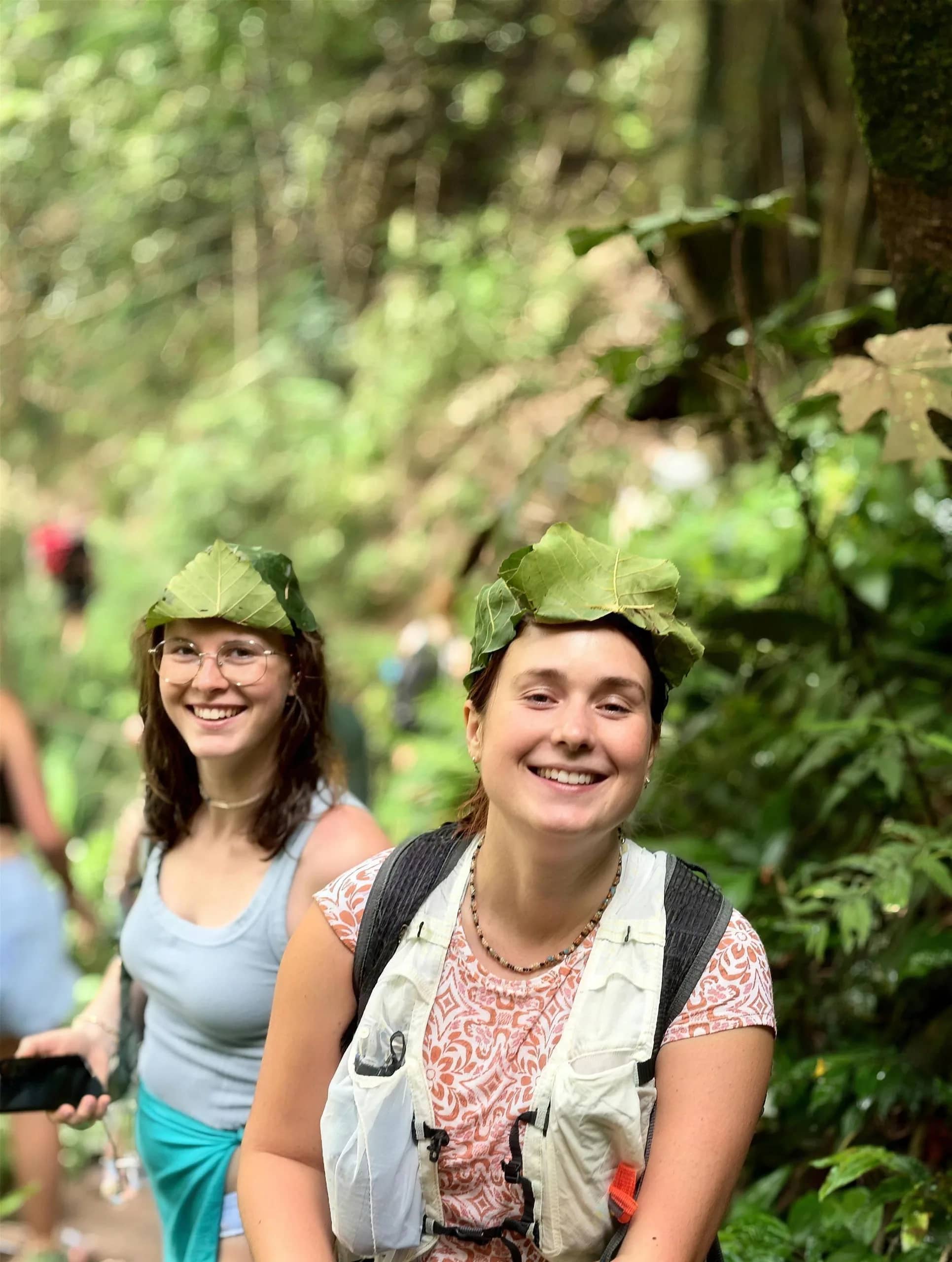 Two smiling hikers wear hats made from leaves on a shaded jungle trail in the Doi Inthanon area