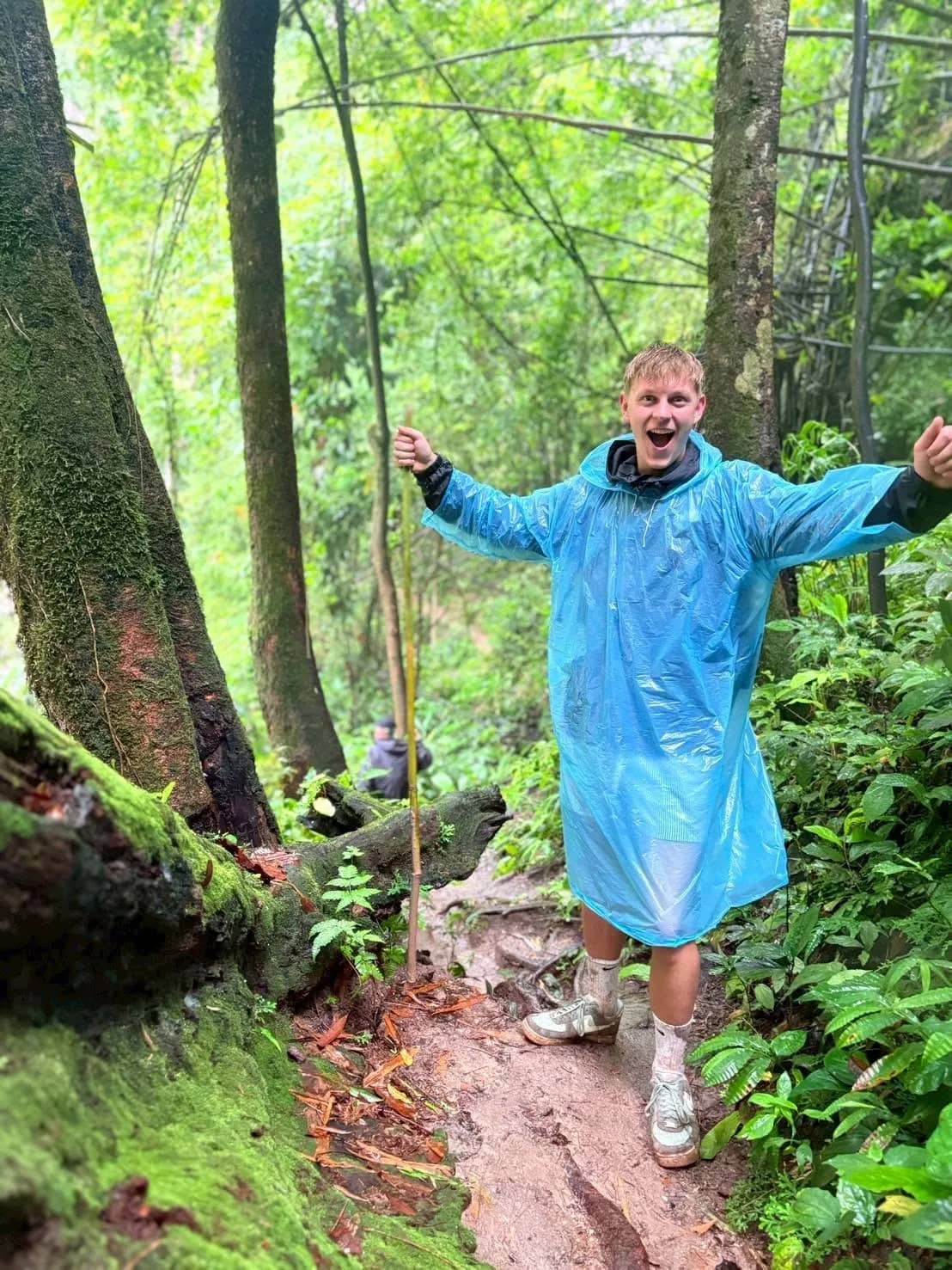 A cheerful hiker in a blue rain poncho stands with arms wide on a muddy jungle path