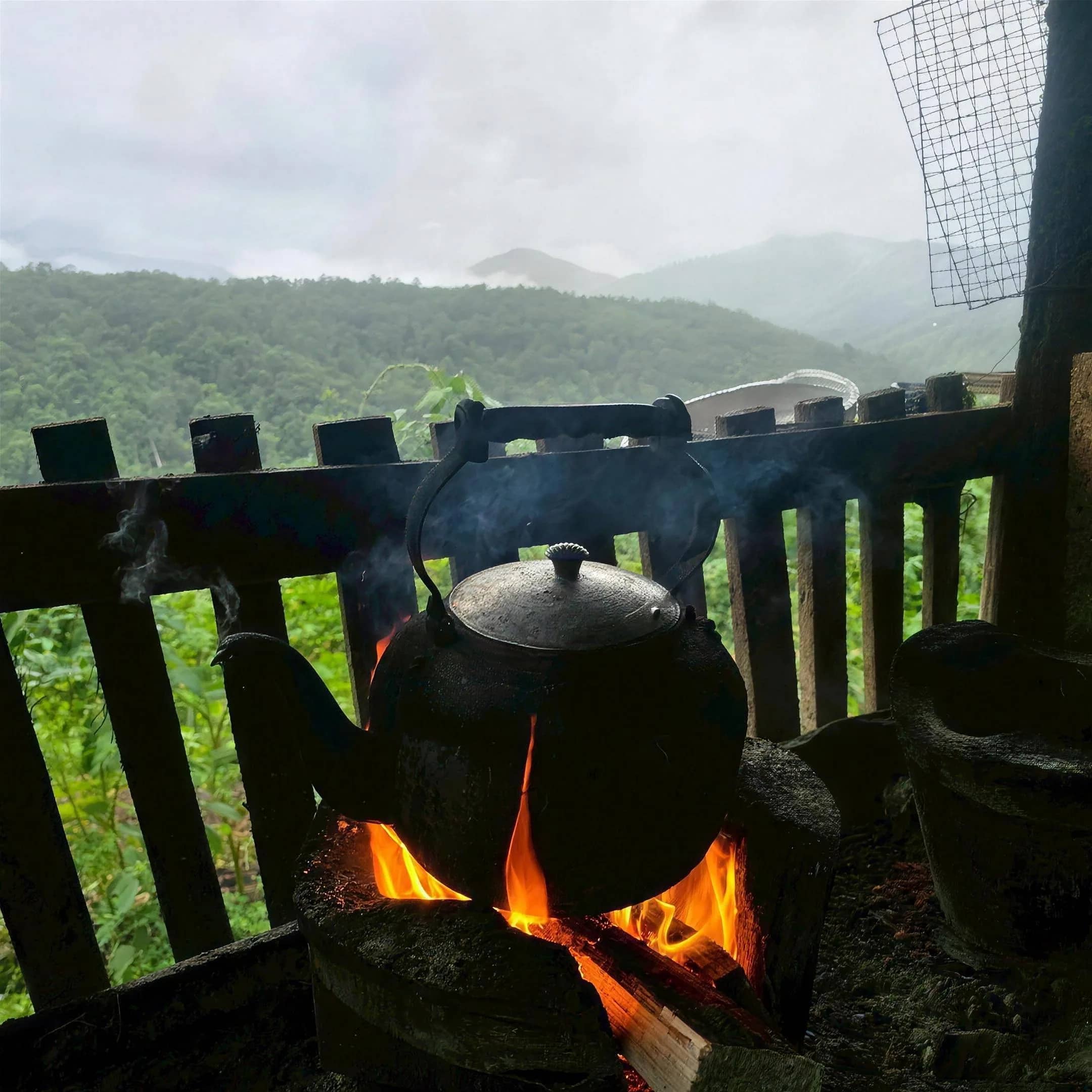 A black kettle heats over a wood fire beside a railing with misty mountain views