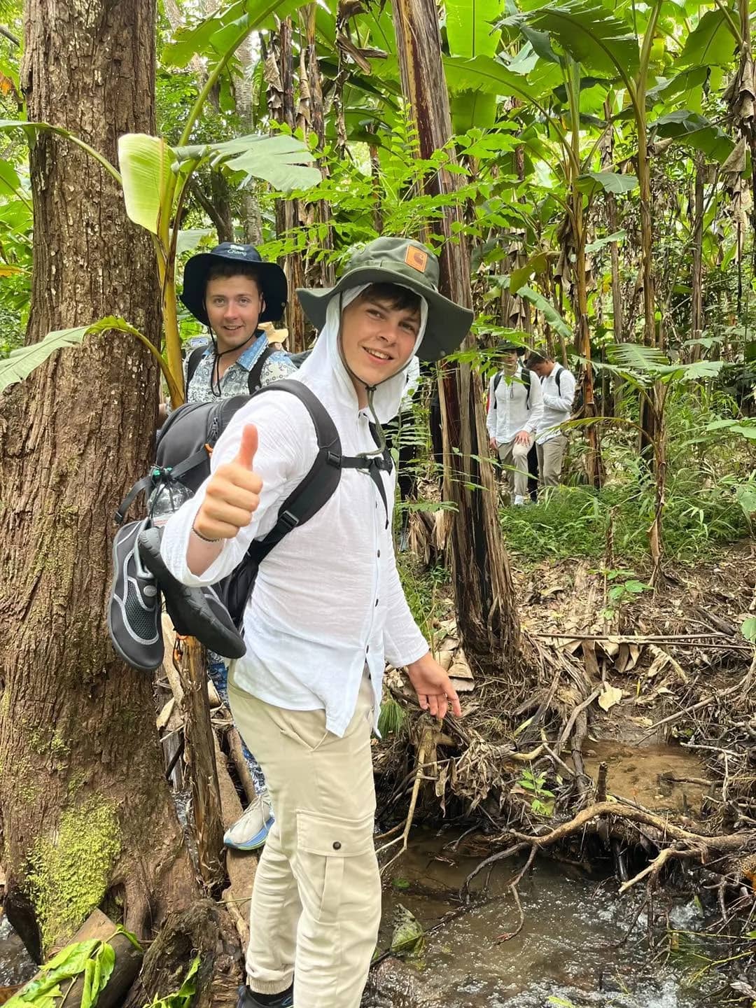 A trekker in a sun hat gives a thumbs up beside a stream on a shady jungle trail