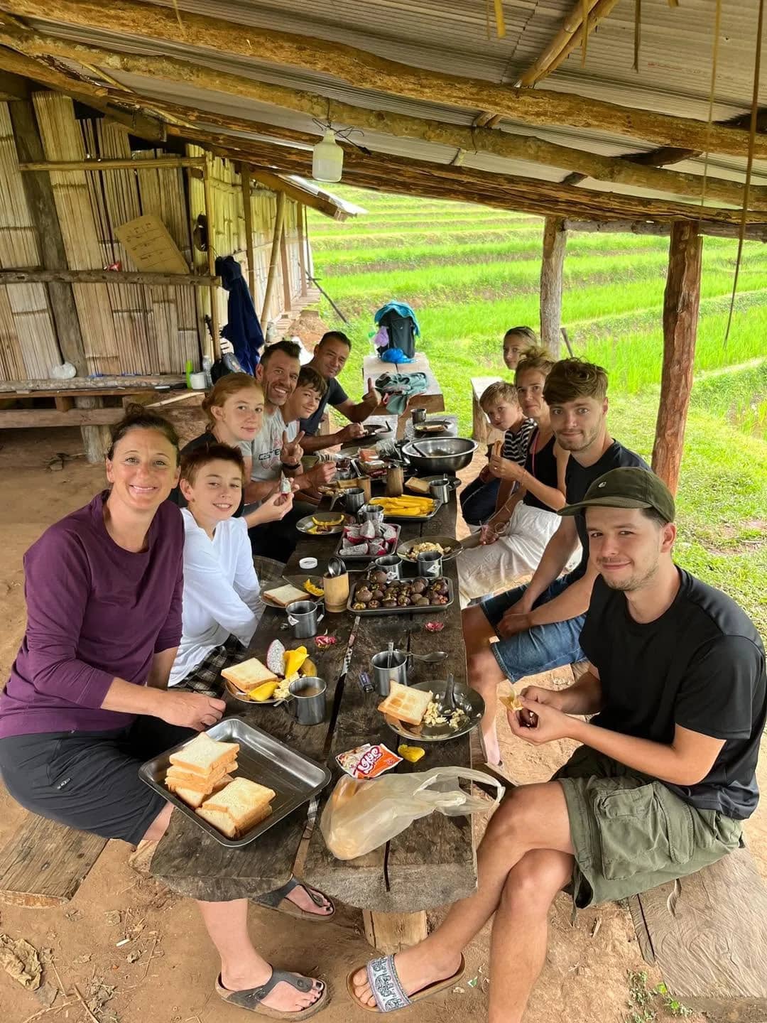 Trekkers share a meal at a long wooden table under a shelter beside green rice fields