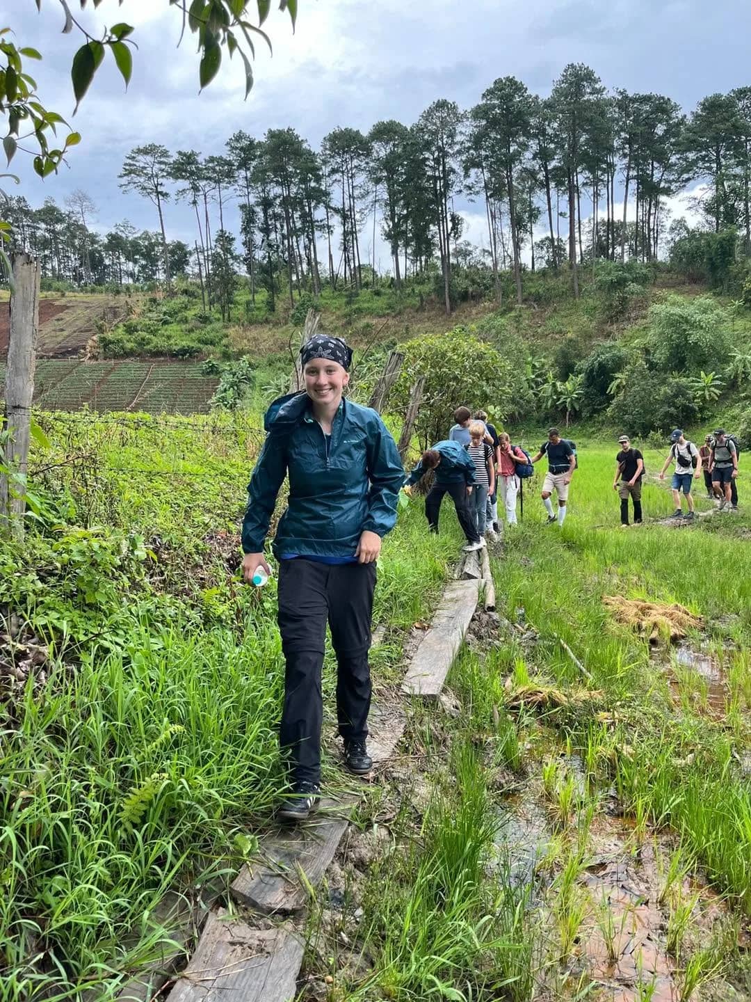 Trekkers in rain jackets walk single file along a narrow path between rice paddies
