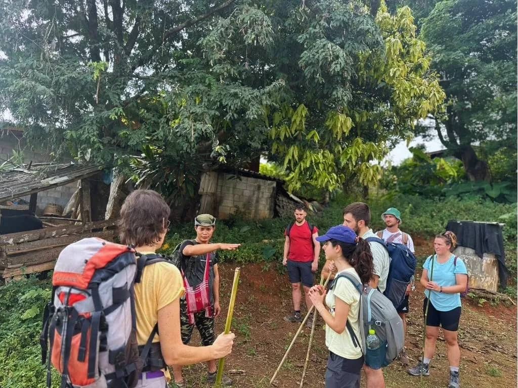 A guide points ahead as trekkers with backpacks and bamboo sticks gather beside a hut.