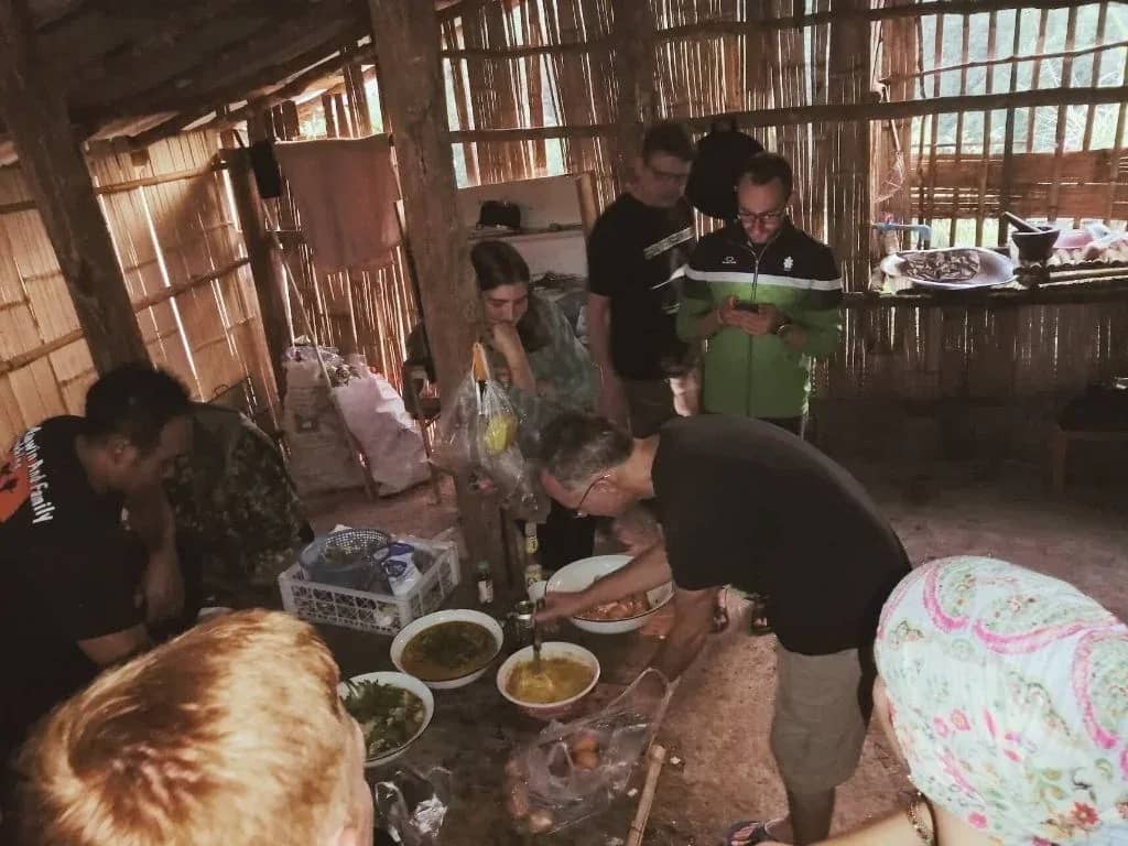 Trekkers and a guide prepare bowls of food together inside a dim bamboo kitchen.