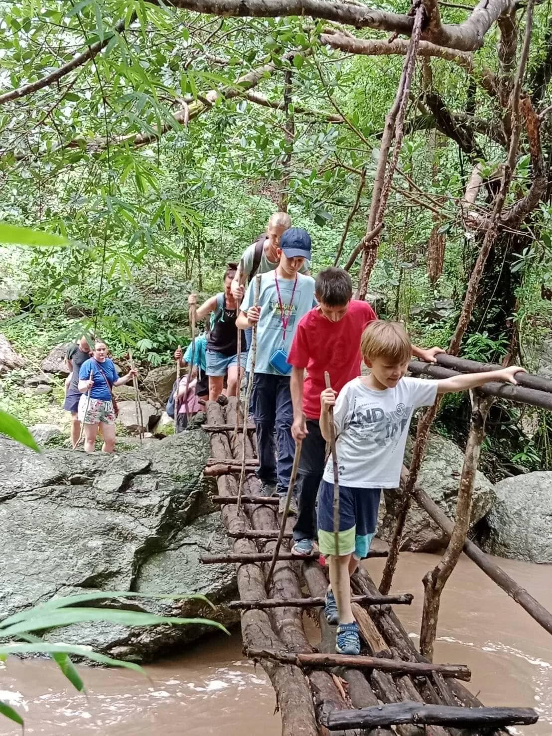 Trekkers and children walk single file across a rustic bamboo bridge over a stream
