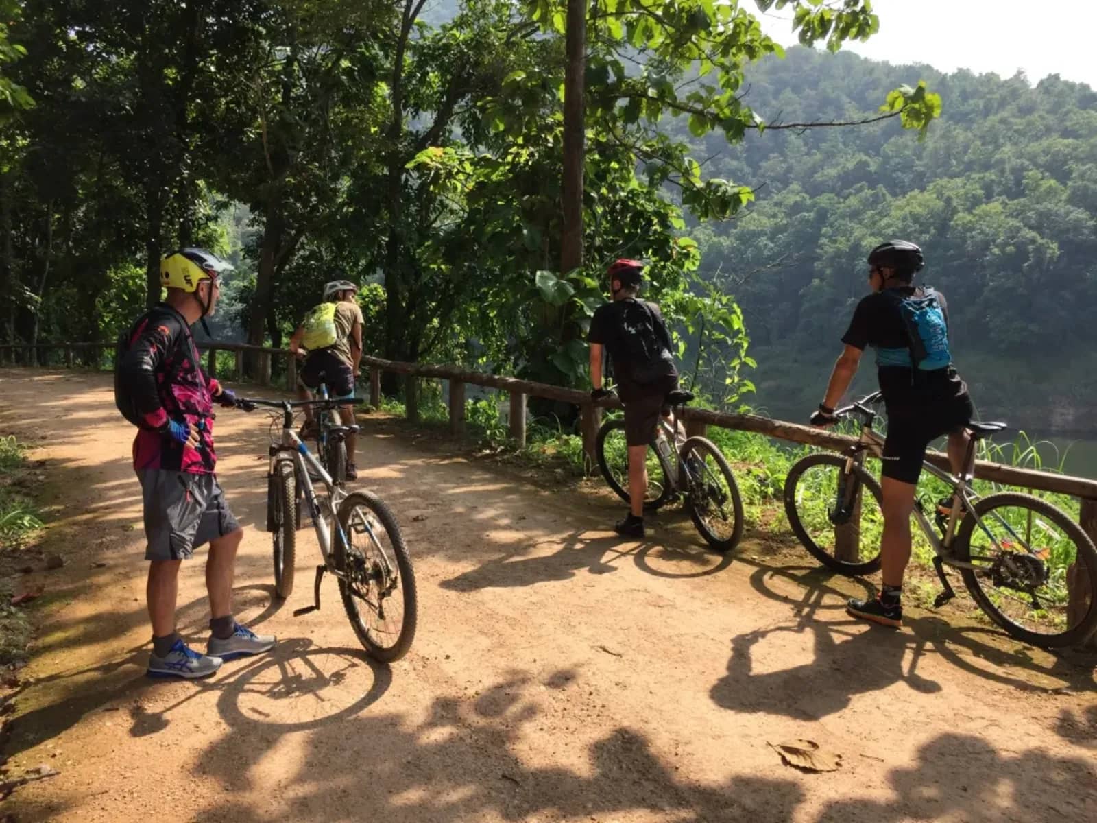 Mountain bikers pause with their bikes on a dirt track beside a lake surrounded by forested hills