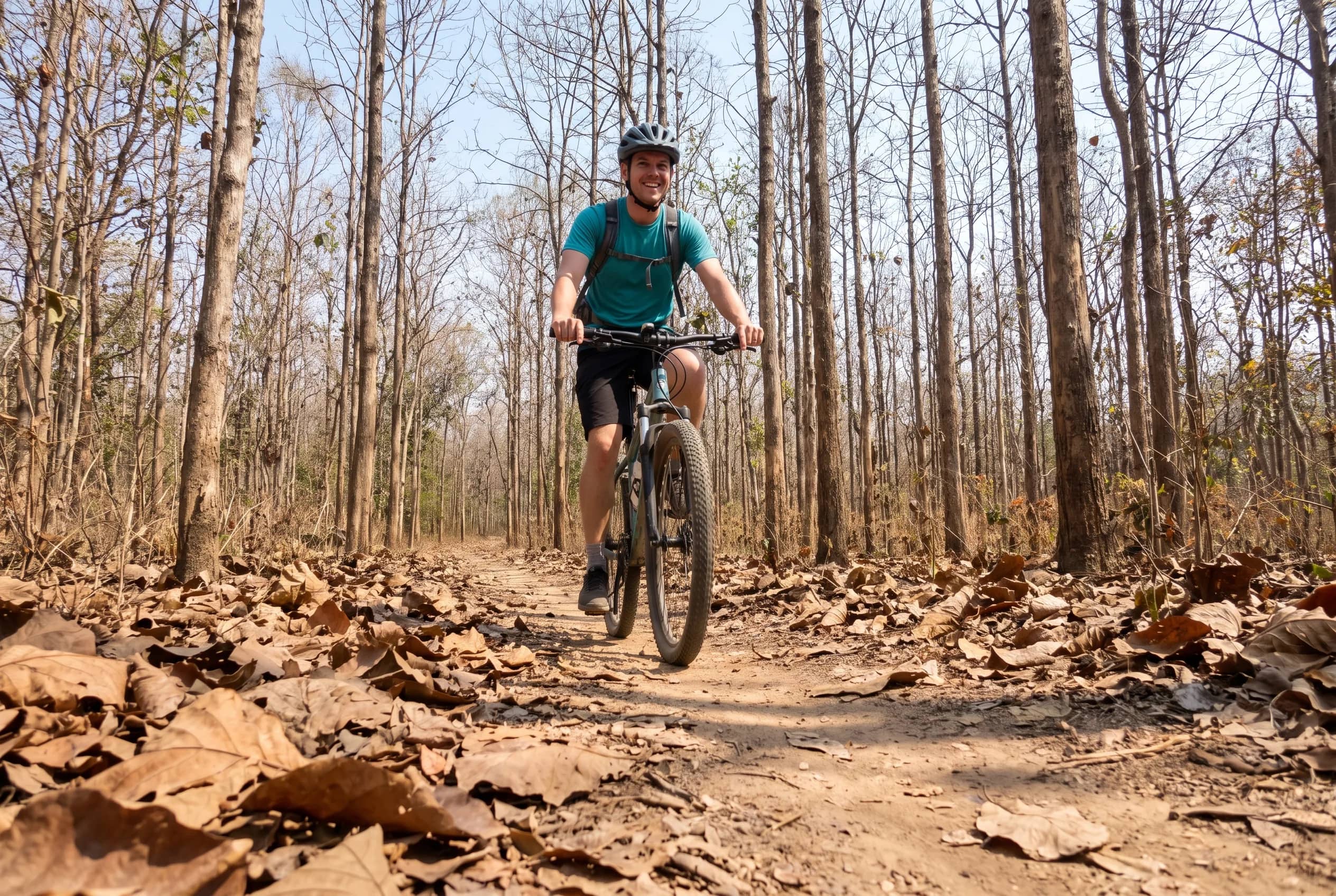 A smiling cyclist in a teal shirt rides a mountain bike along a leaf-covered trail in dry forest