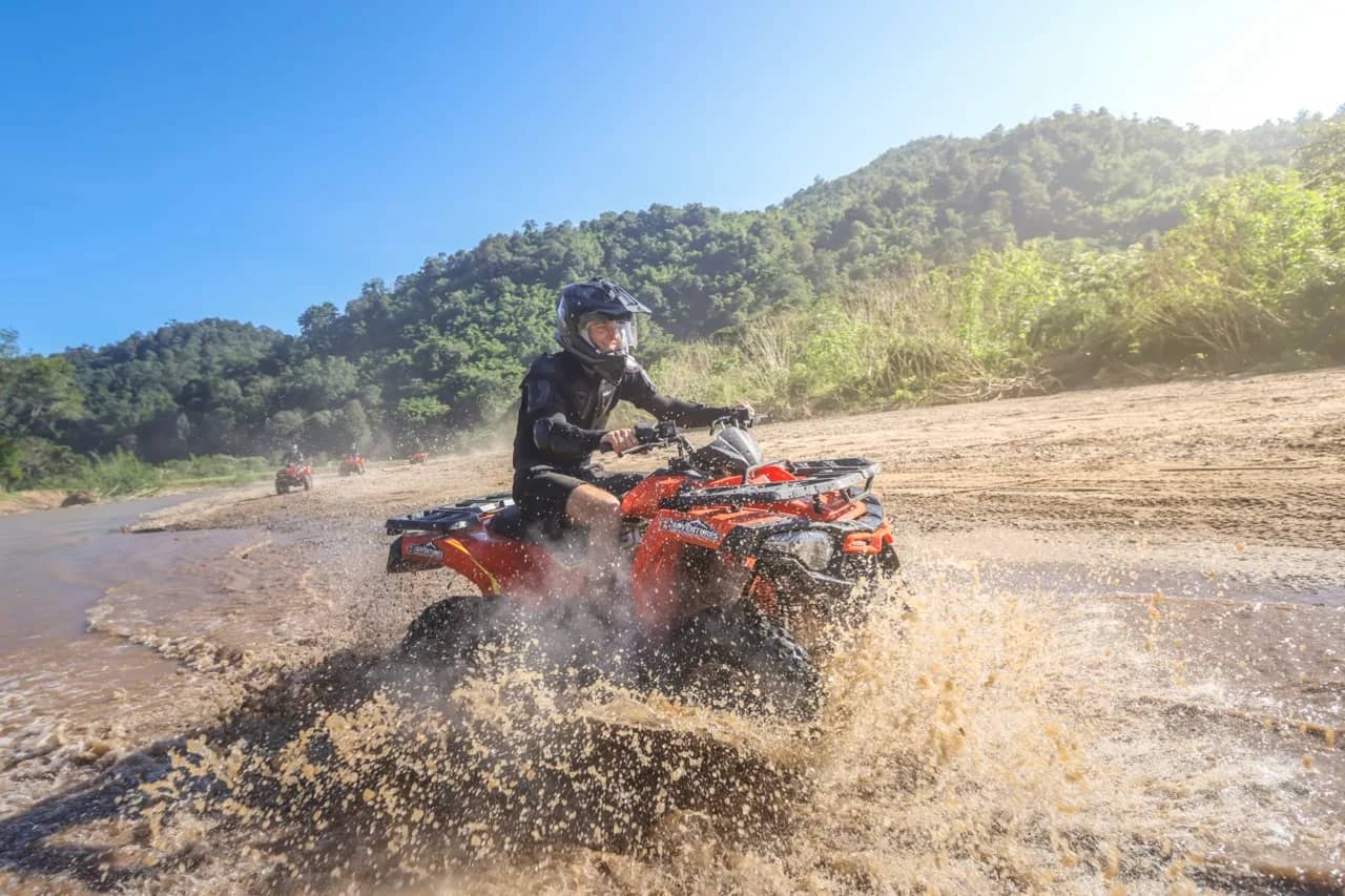 A rider in a helmet drives an orange ATV through shallow river water sending up spray