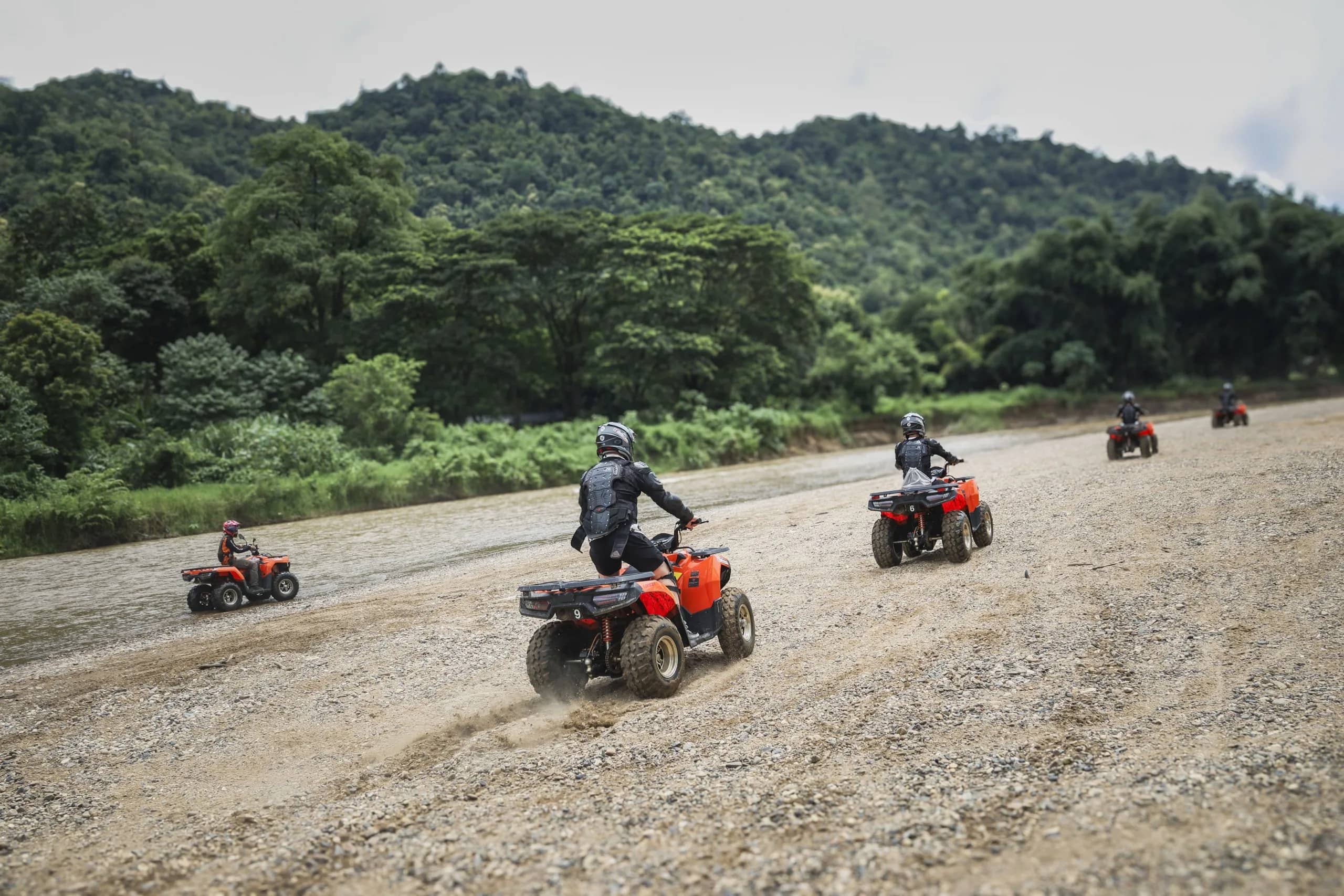 A line of ATVs drives across a wide gravel riverbed below forested hills