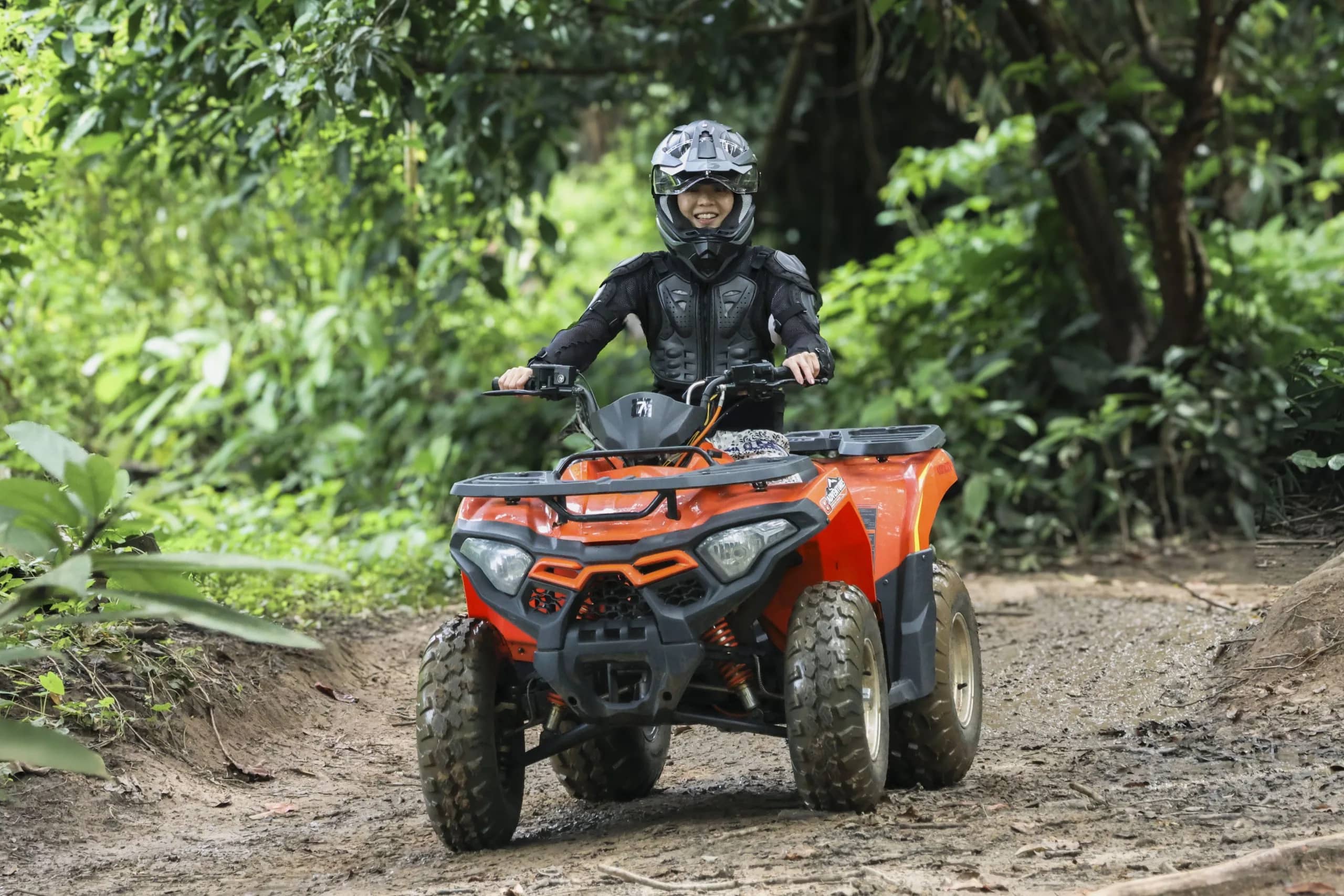A rider in a helmet and armour drives an orange ATV along a muddy jungle track