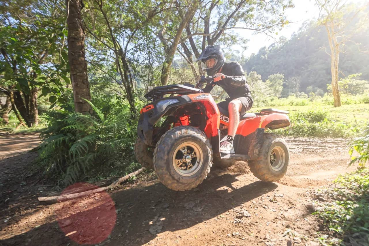 A helmeted rider steers a red ATV up a dirt trail through sunlit jungle