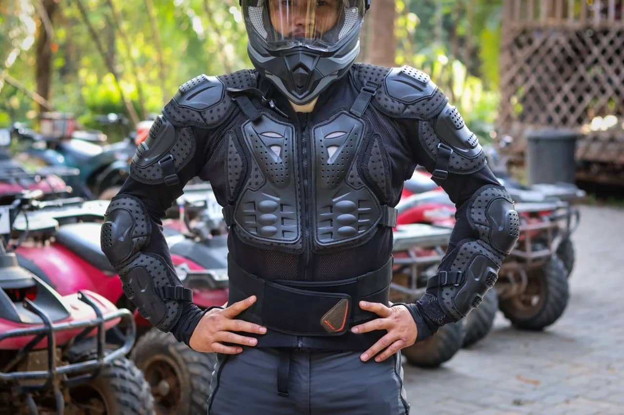 A rider in a full-face helmet and black body armour stands in front of parked ATVs