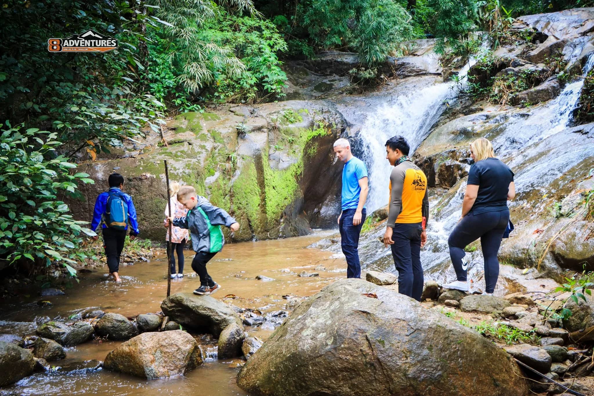A small group of hikers crosses rocks and a shallow stream below a jungle waterfall near Chiang Mai