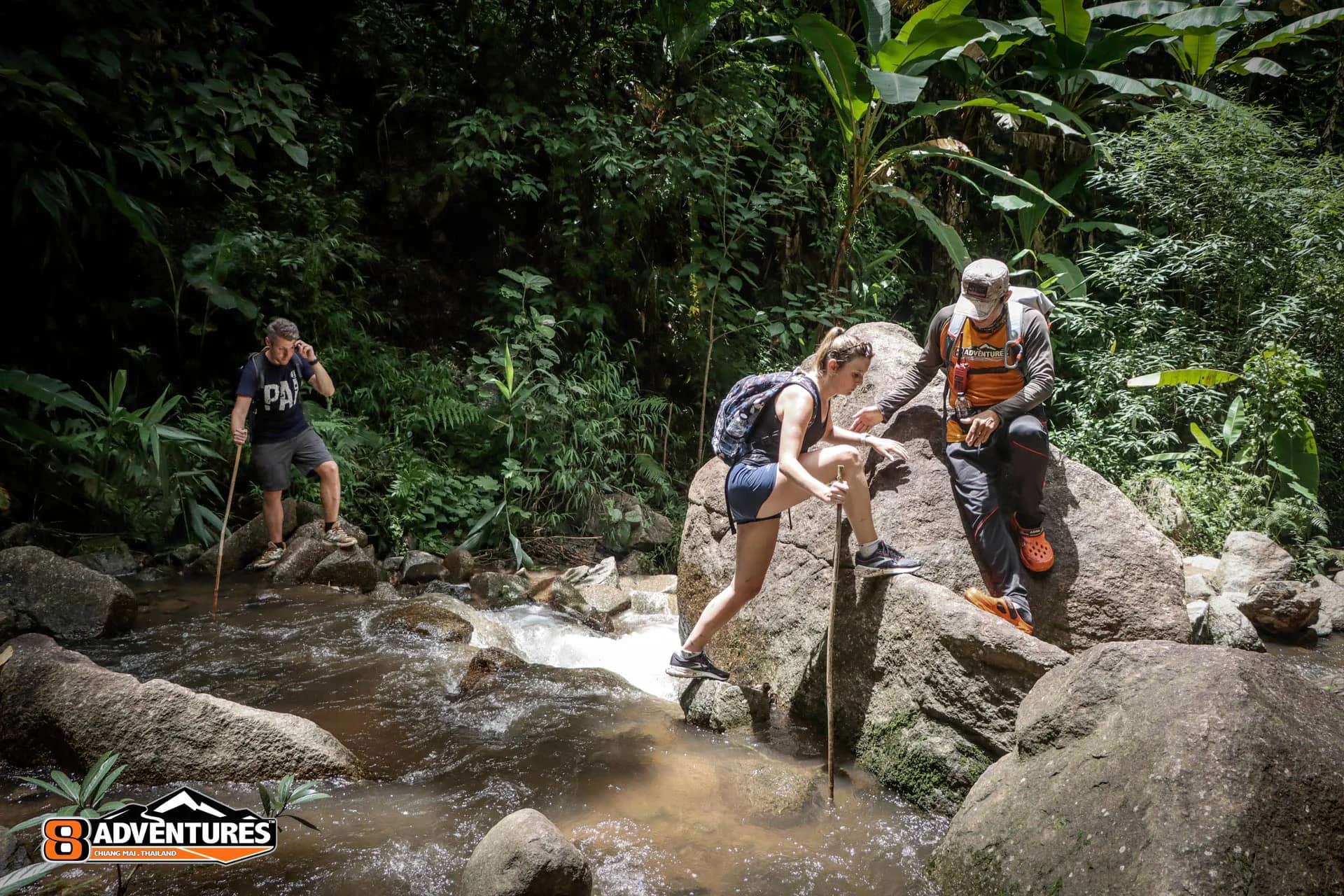 A guide helps hikers with walking sticks cross the boulders of a jungle stream
