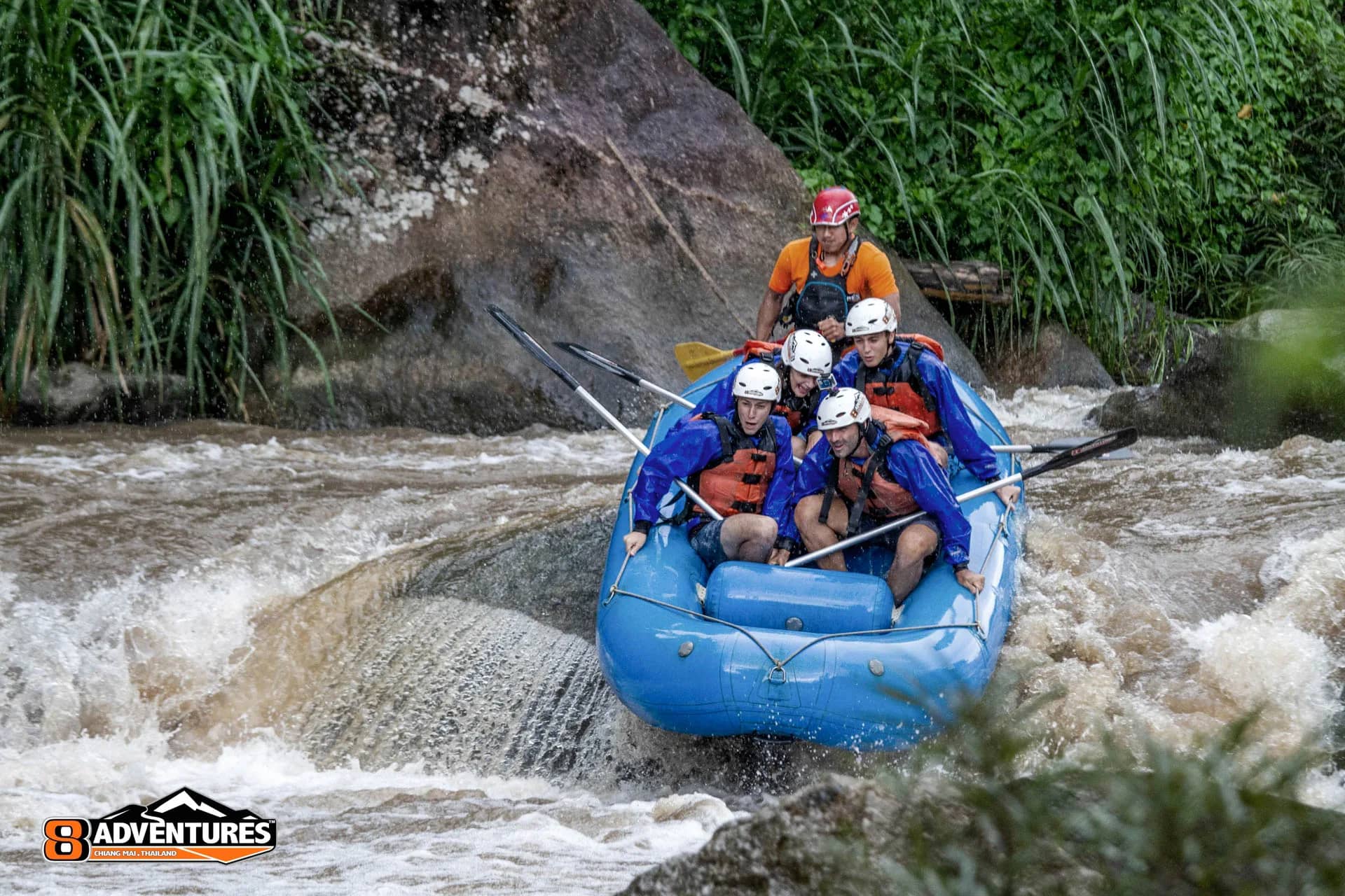 A blue raft of helmeted paddlers drops down a rapid between rocks on the Mae Taeng River