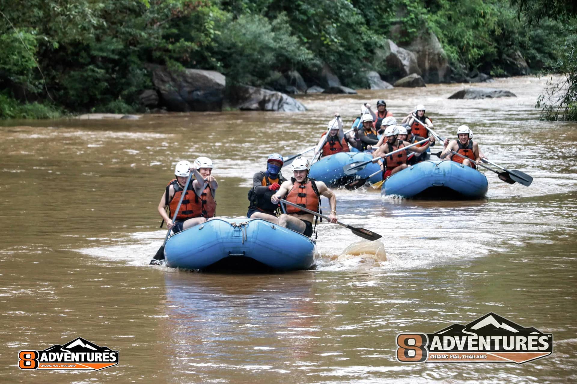A line of blue rafts with paddlers in helmets moves down a wide jungle river