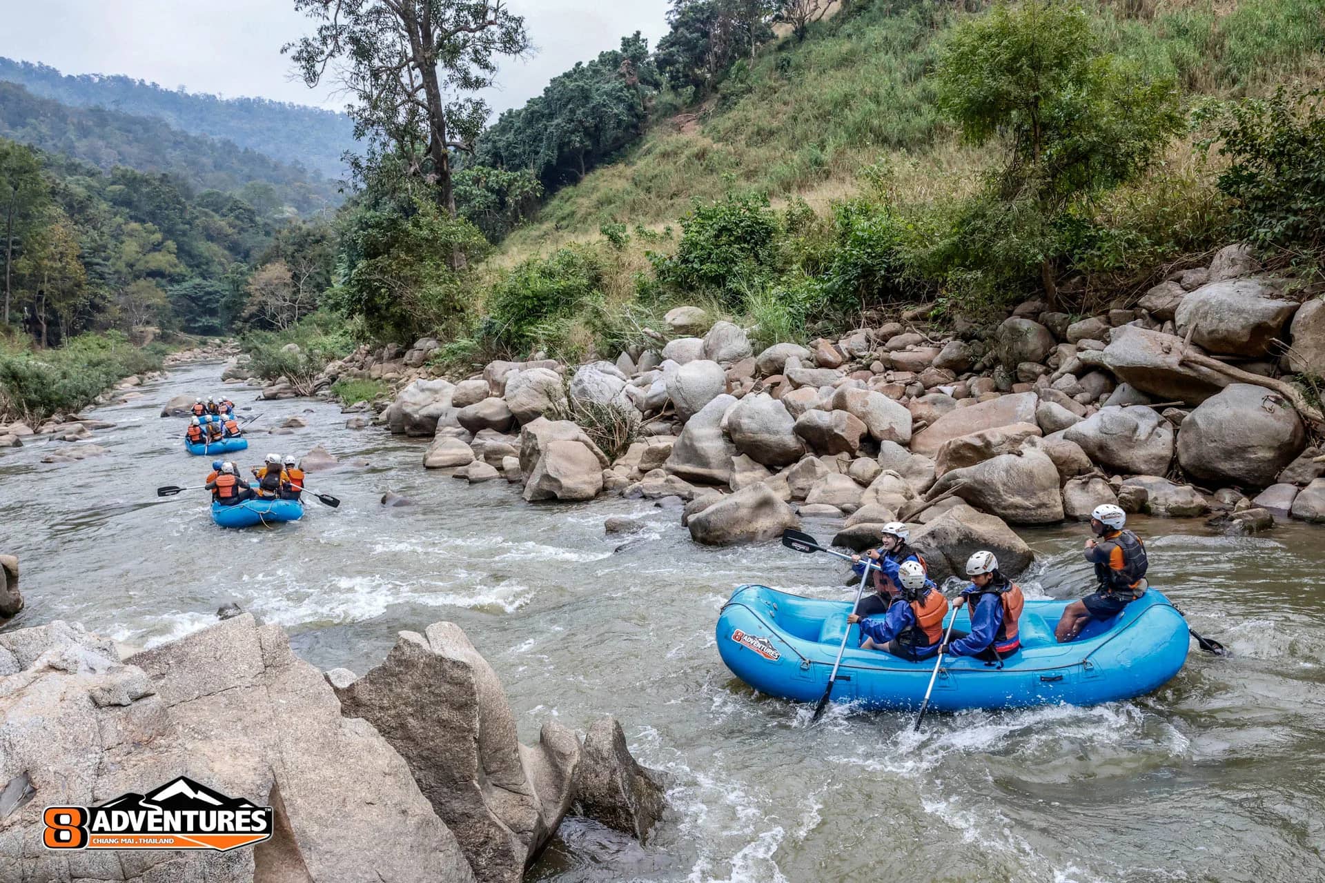Several blue rafts float down a rocky river through a green mountain valley