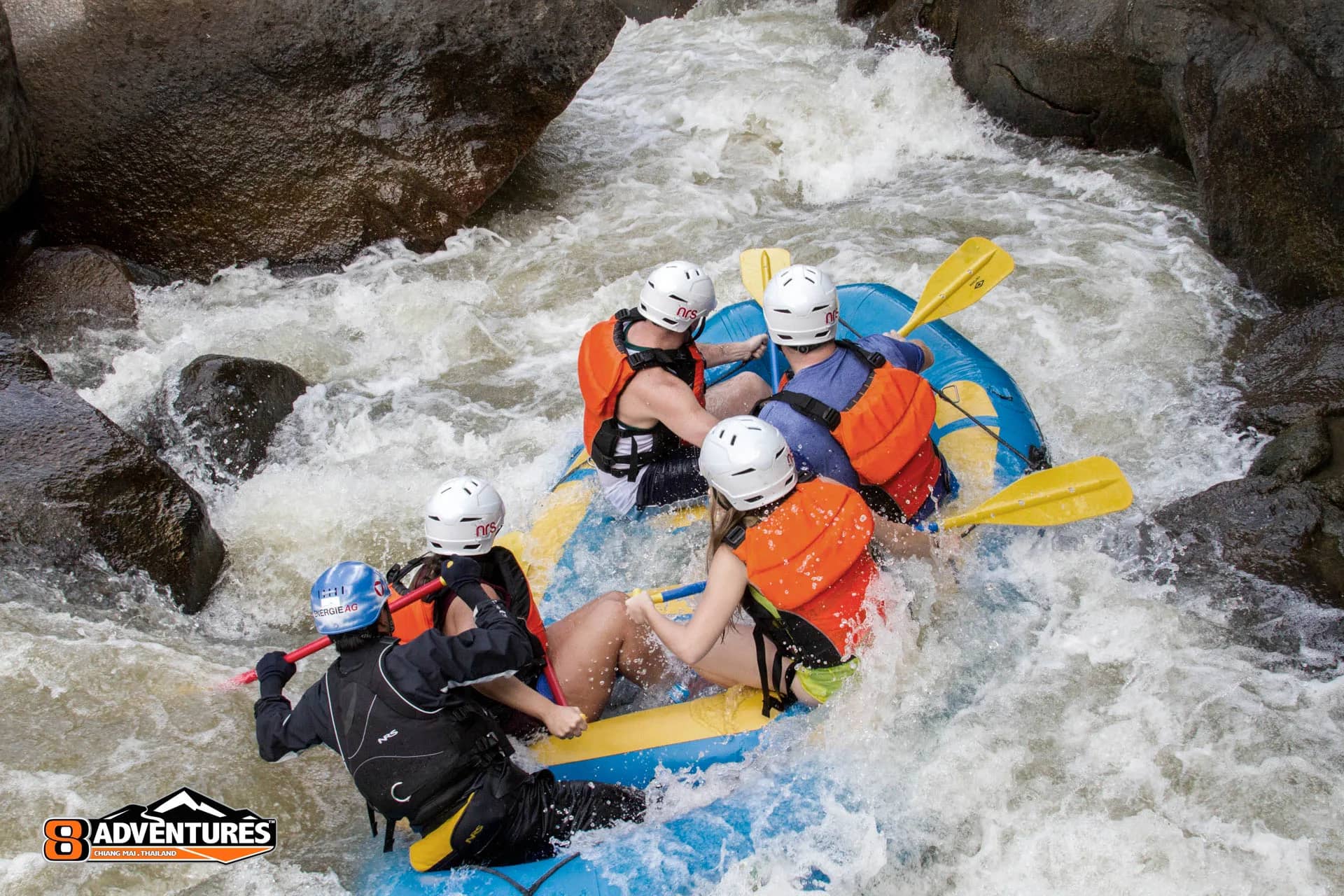 A guide steers a blue raft of helmeted paddlers through whitewater between dark boulders