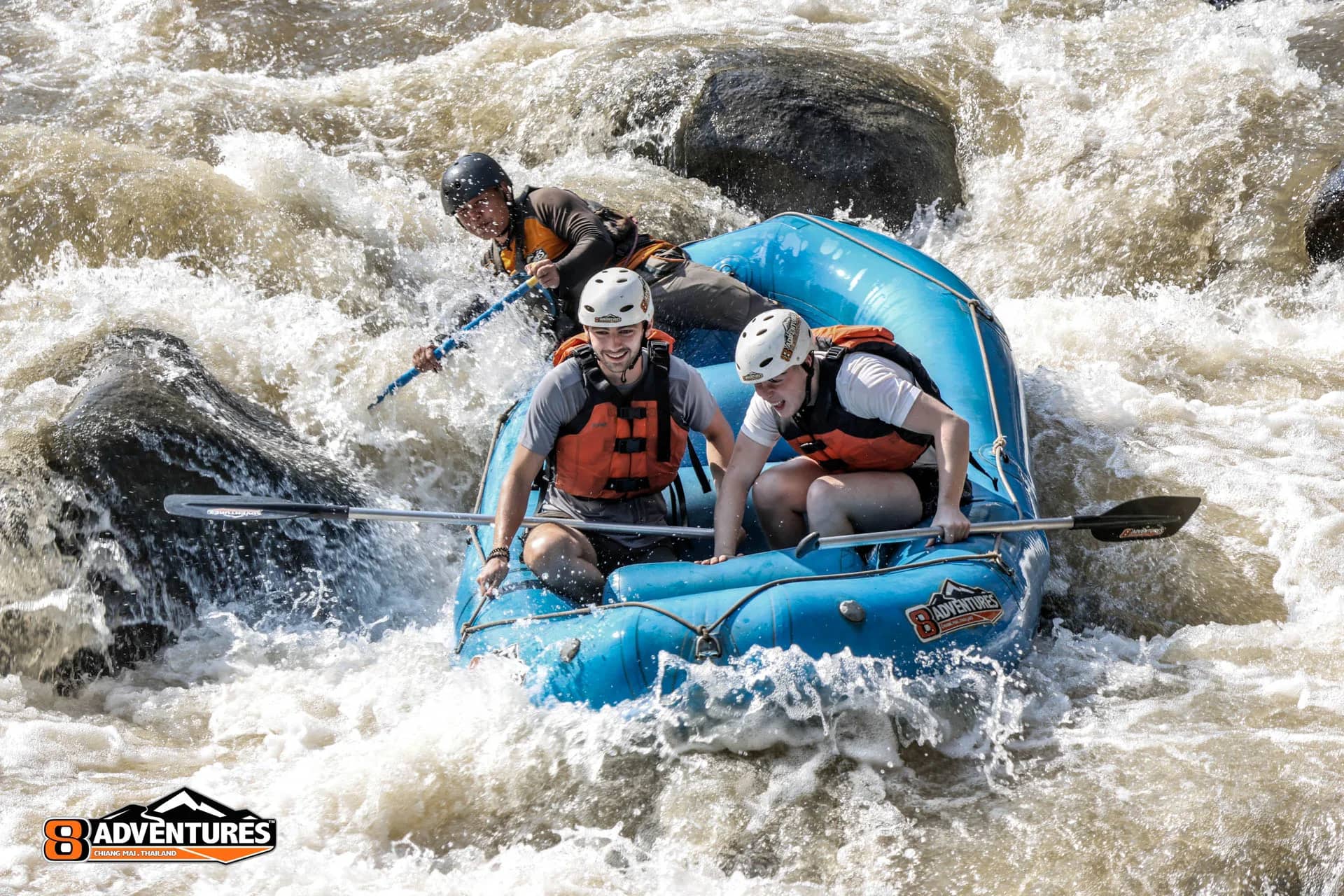 A guide at the back steers a blue raft carrying helmeted rafters through foaming rapids
