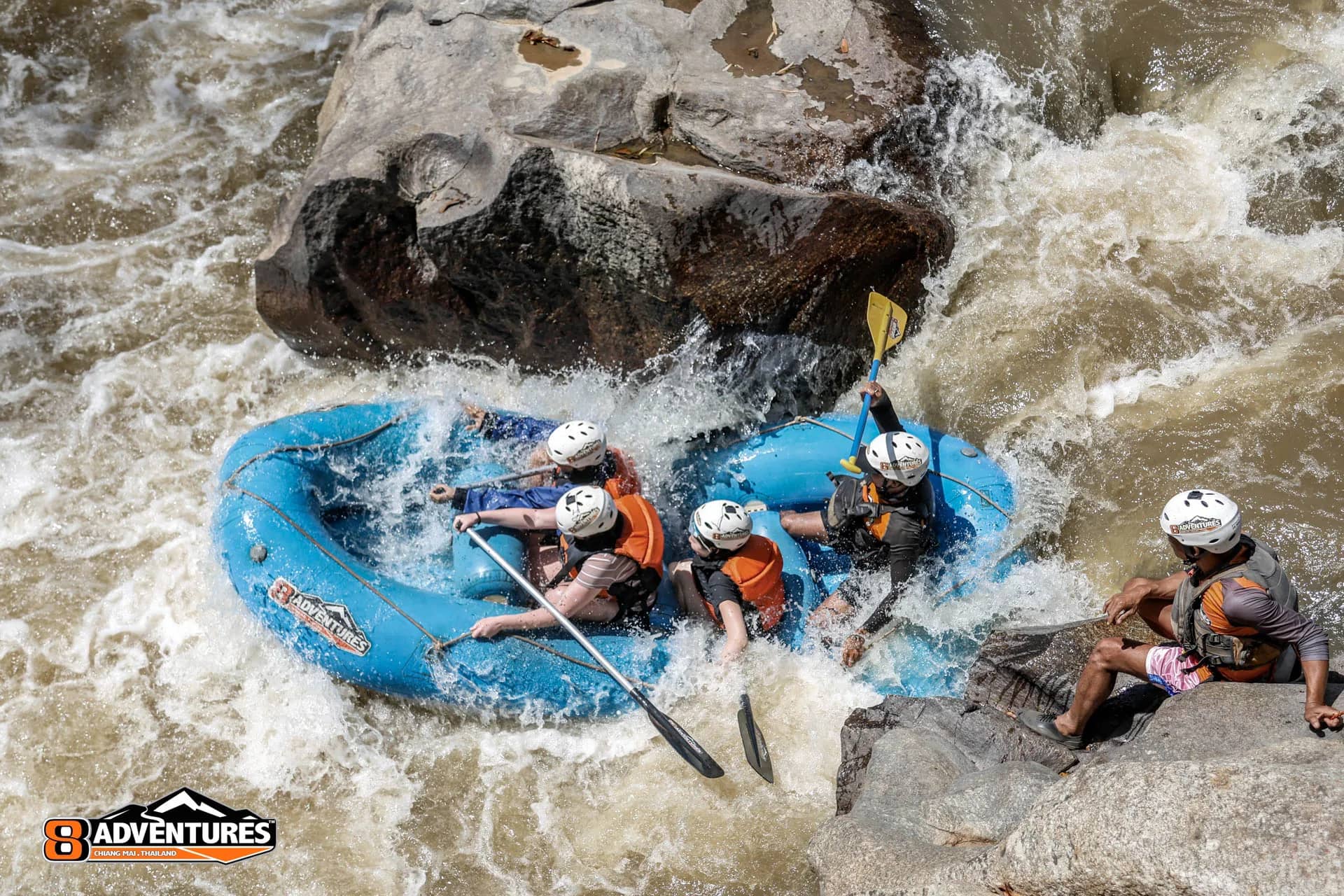 A blue raft slams into rocks in fast whitewater as a guide watches from a boulder