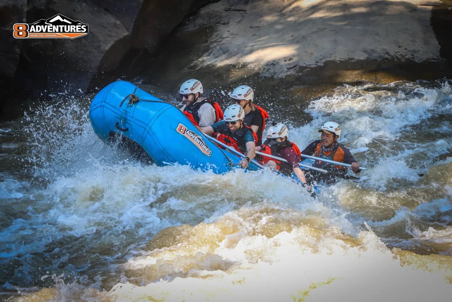 A blue raft with helmeted paddlers tilts through foaming rapids between large boulders
