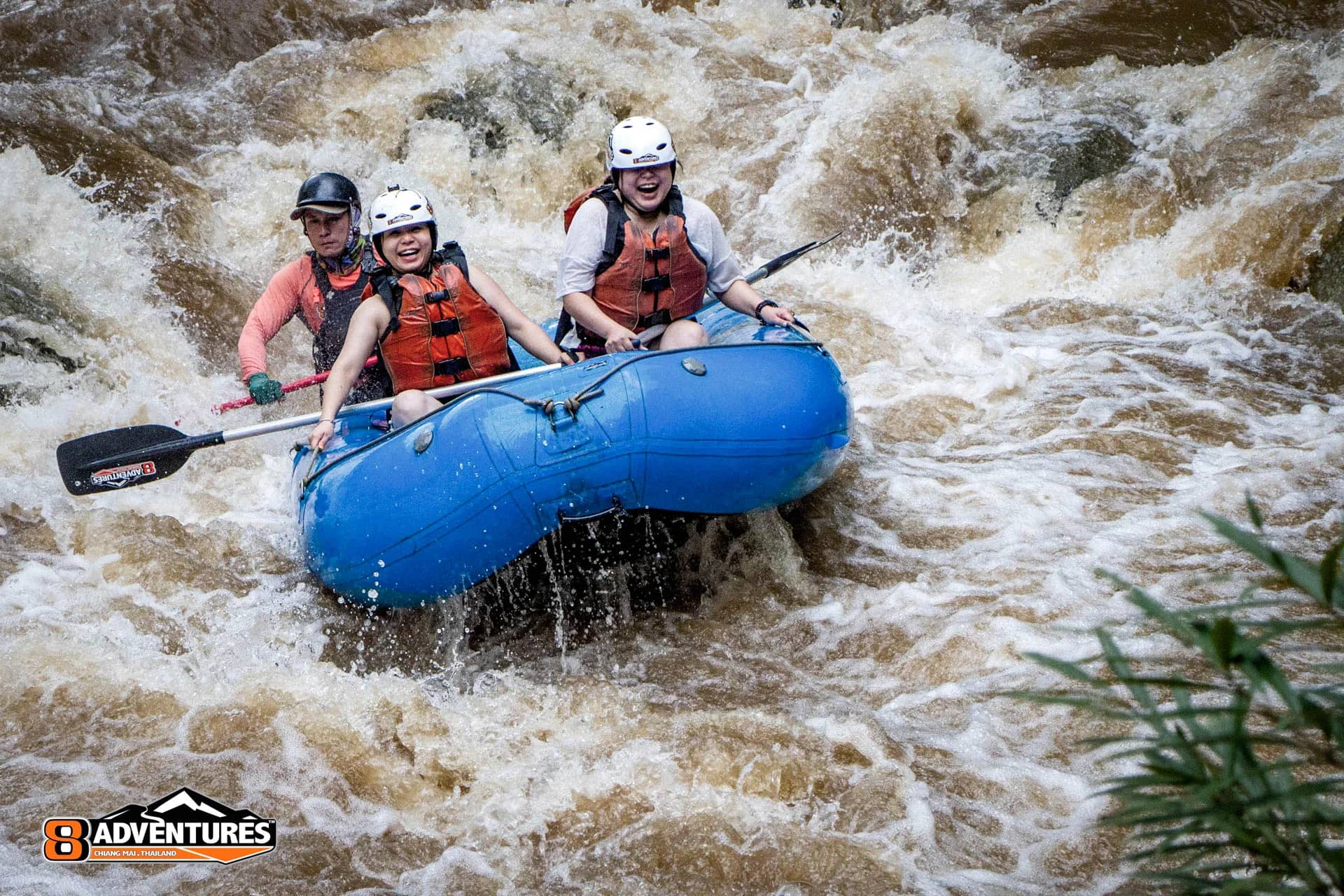 Rafters in helmets and life jackets laugh as their blue raft crashes through brown whitewater