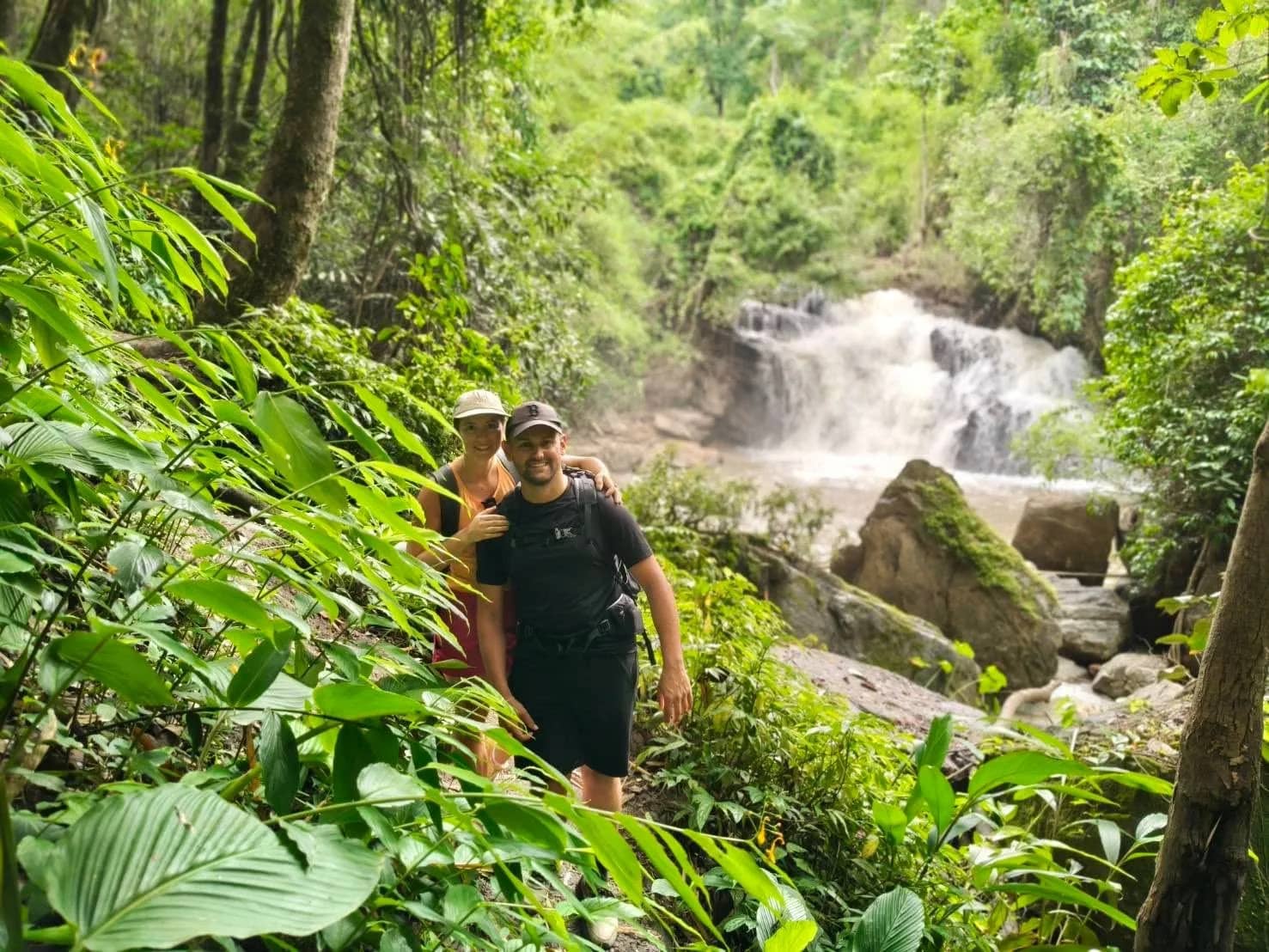 Two hikers walk through lush jungle undergrowth in front of a rocky waterfall.