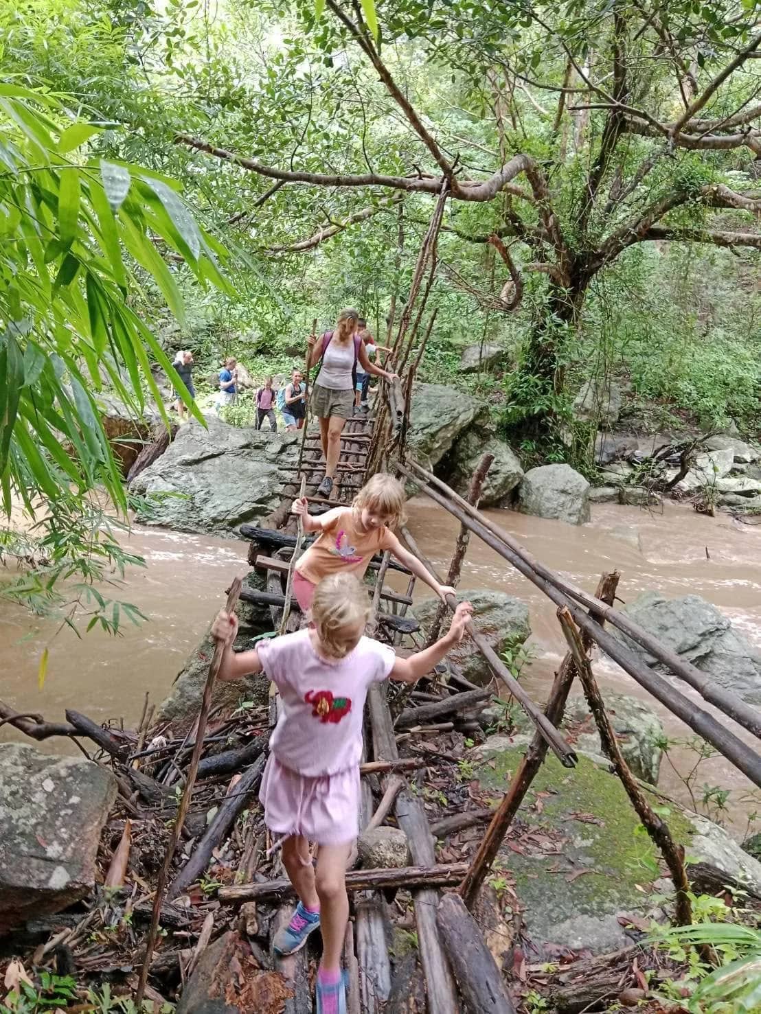 Children cross a rickety bamboo bridge over a brown jungle river as the group follows.