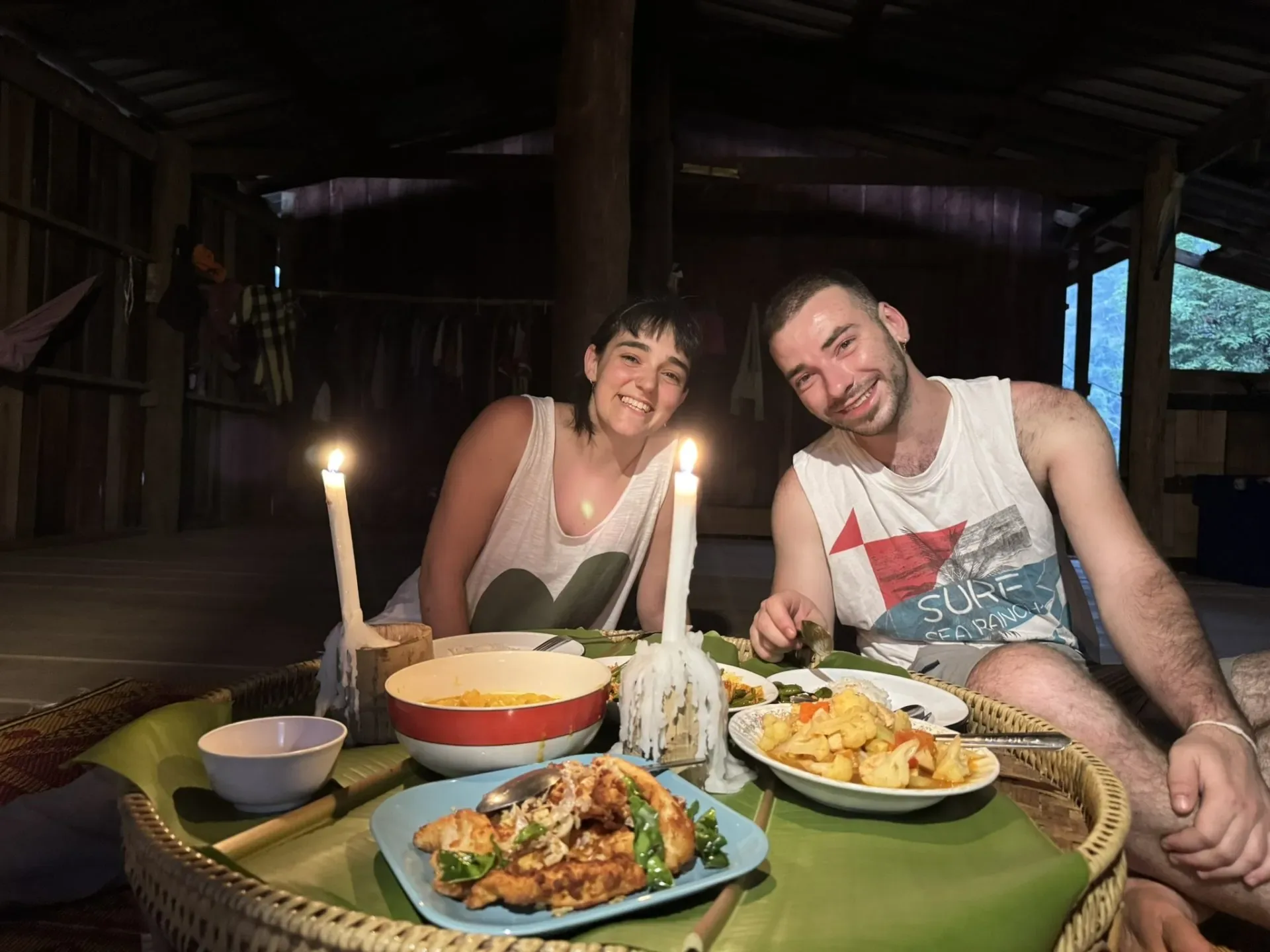 Two trekkers smile over a candlelit dinner of Thai dishes on a rattan tray in a wooden hut