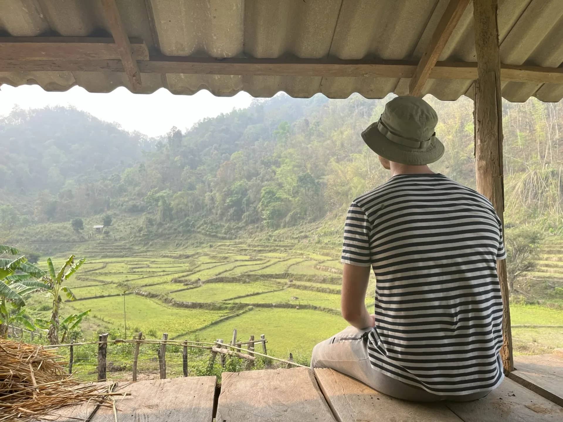 A trekker in a bucket hat sits under a shelter gazing at rice terraces and forested hills