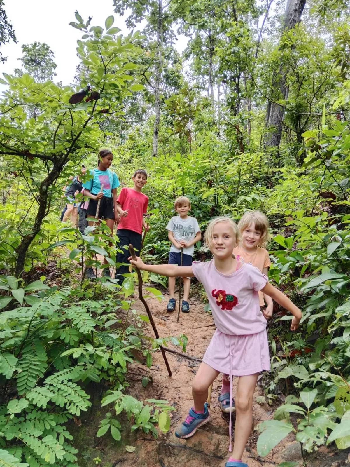 Children with walking sticks lead a family up a narrow trail through green jungle