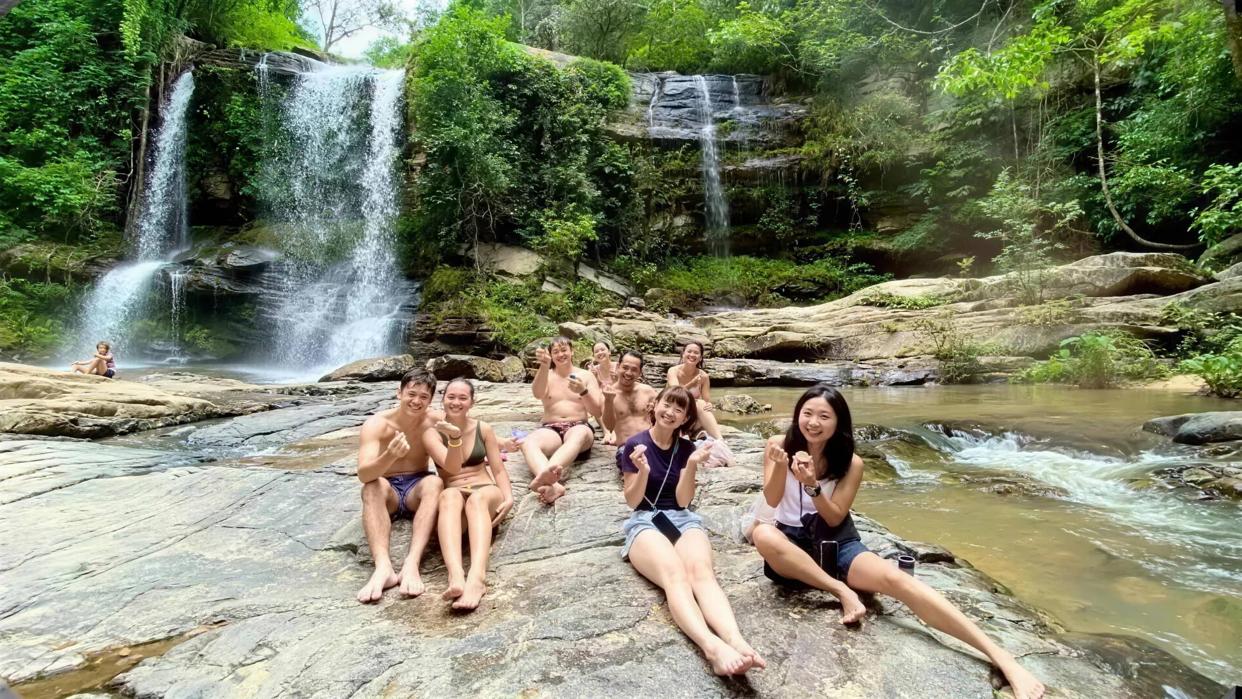 Visitors sit on smooth rocks in front of a tiered waterfall in green jungle