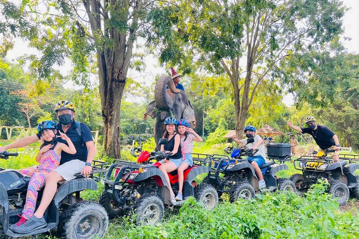 Riders including children sit on ATVs lined up on a grassy trail with an elephant behind them