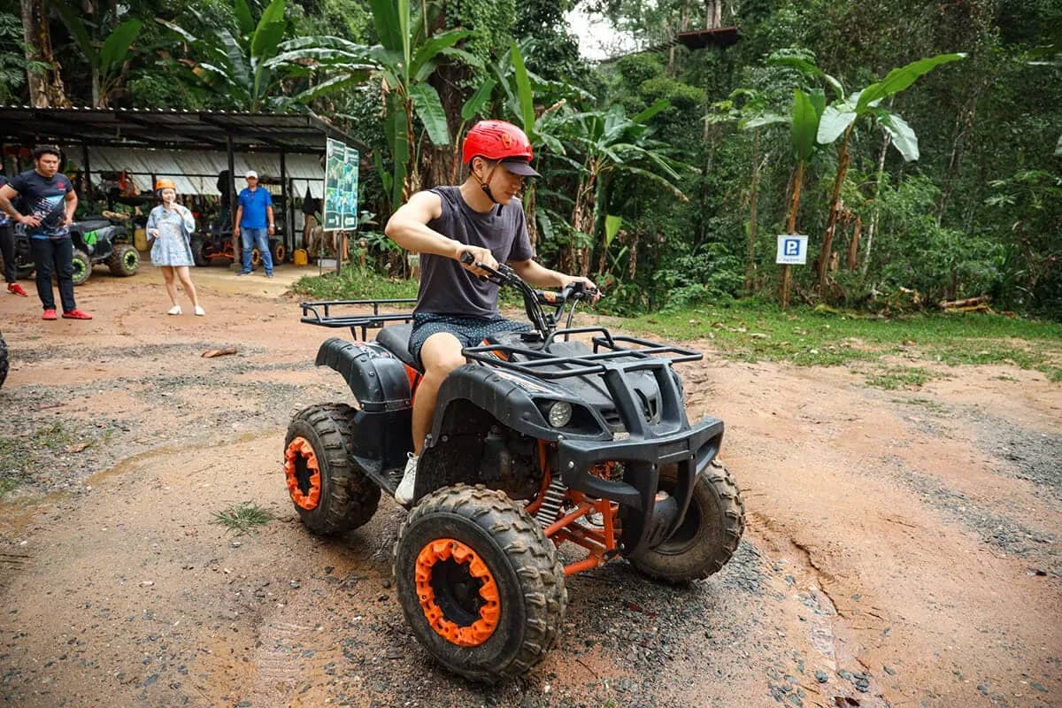A rider in a red helmet drives a small ATV across a dirt yard in front of the base camp