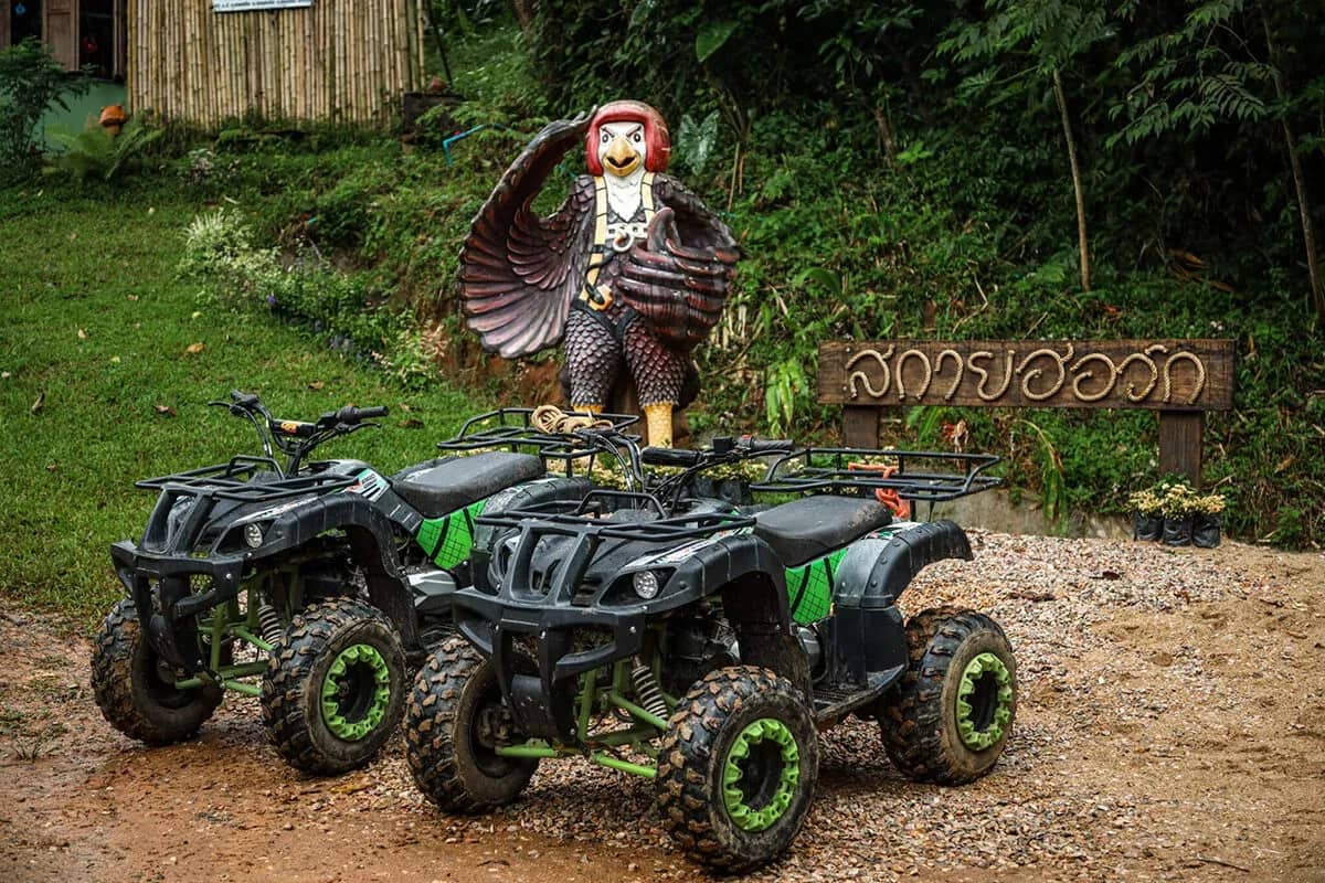 Two black and green ATVs stand on a dirt clearing beside a carved figure and a wooden sign