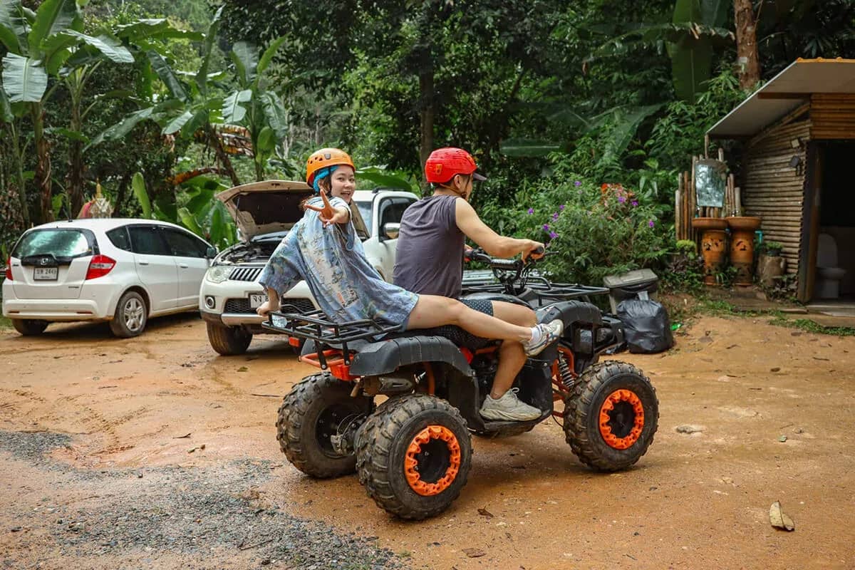 Two riders in helmets sit on an orange-wheeled ATV on a red dirt yard beside parked cars