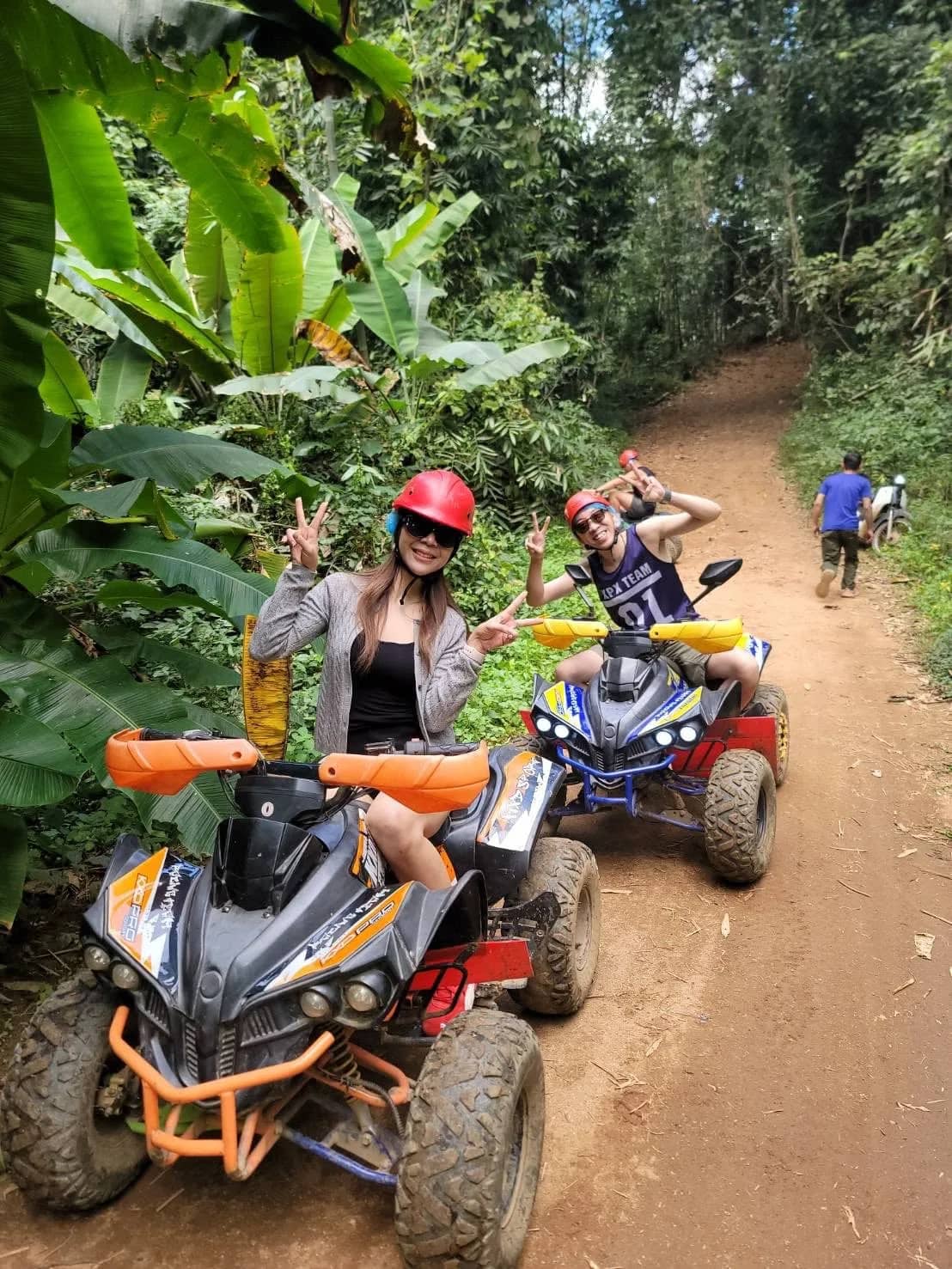 Two riders flash peace signs from their ATVs on a narrow dirt track through banana trees