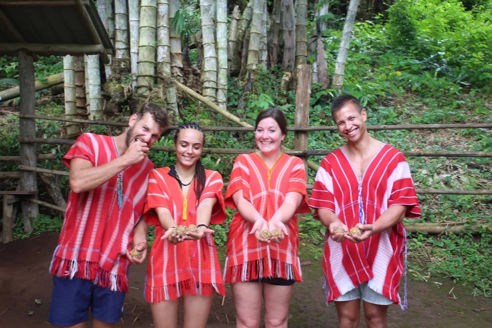 Four visitors in red tunics hold out small herbal balls in their hands beside a bamboo fence