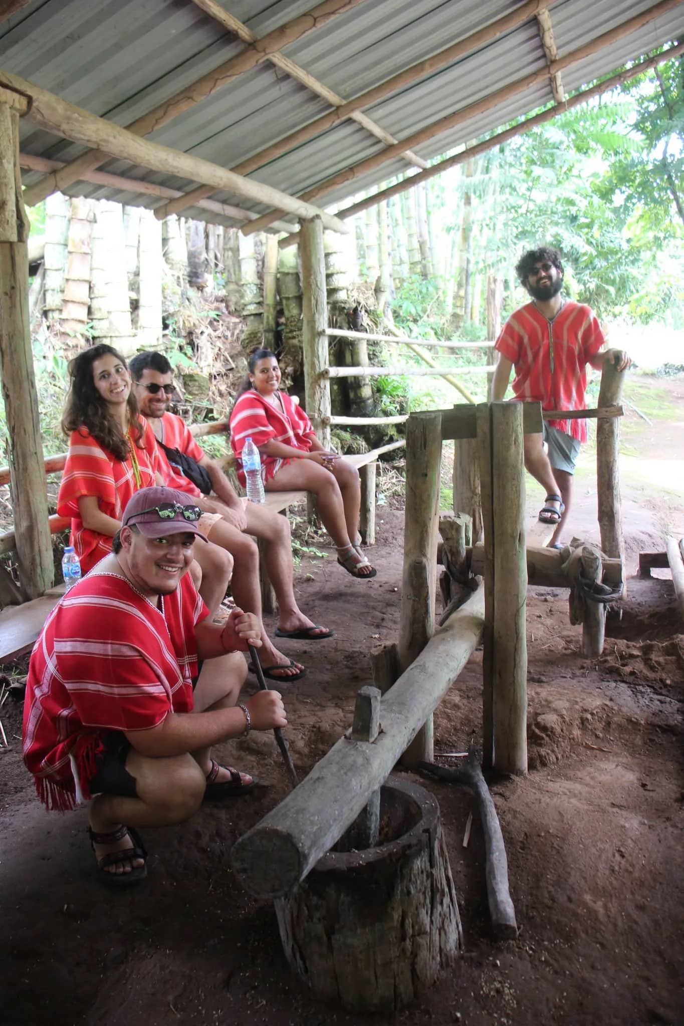 Visitors in red Karen shirts pound food in a wooden mortar under a shelter at the elephant camp